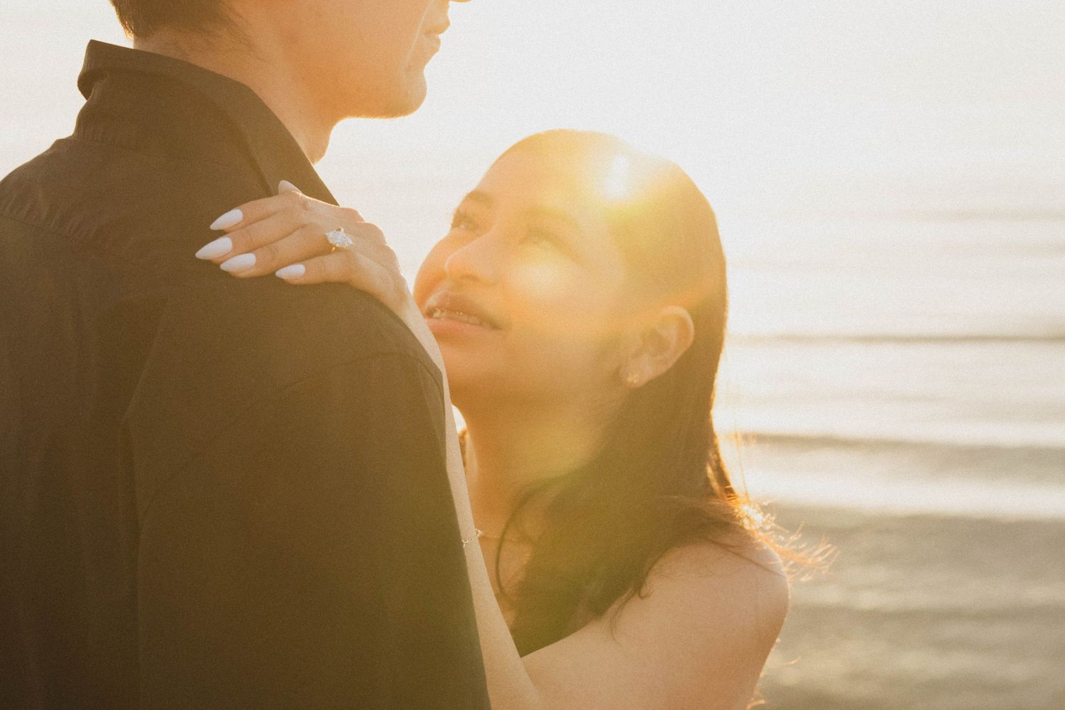Romantic couple holding each other close at sunset by the ocean with golden lens flare.