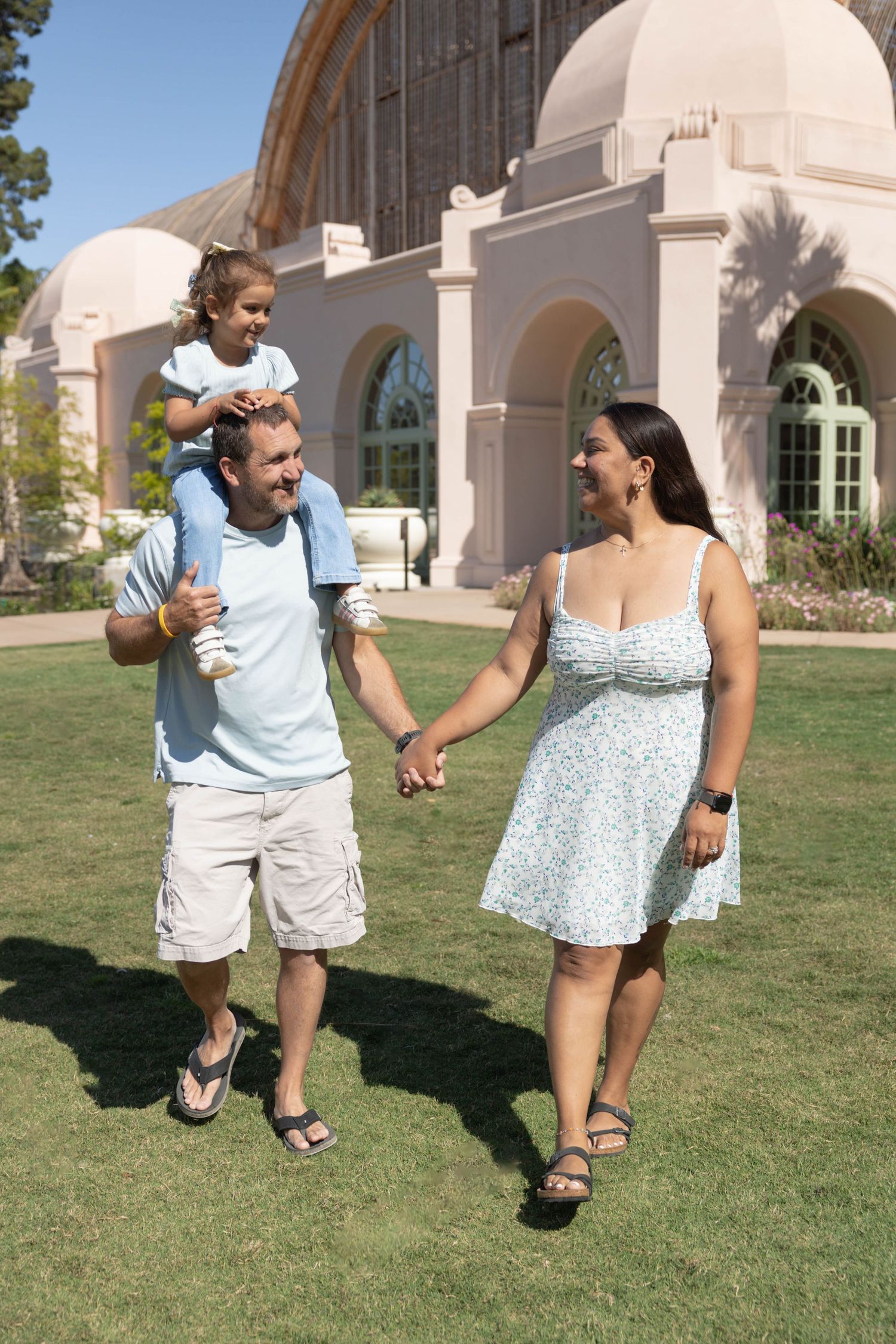 Happy family walking outdoors, father carrying daughter on shoulders, holding hands with smiling mother near elegant building.