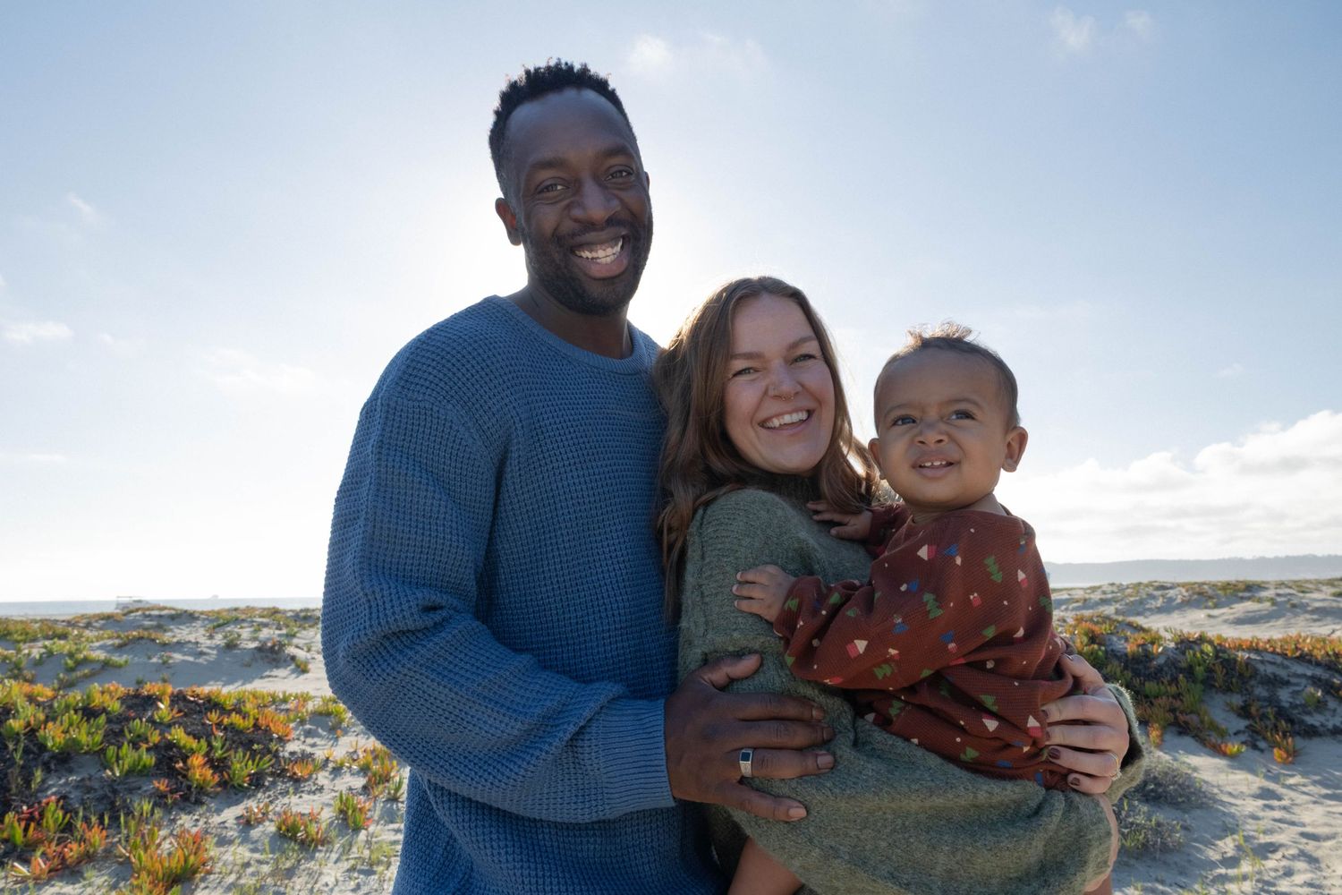 Happy interracial family with baby smiling together on a sunny beach with dunes and blue sky.