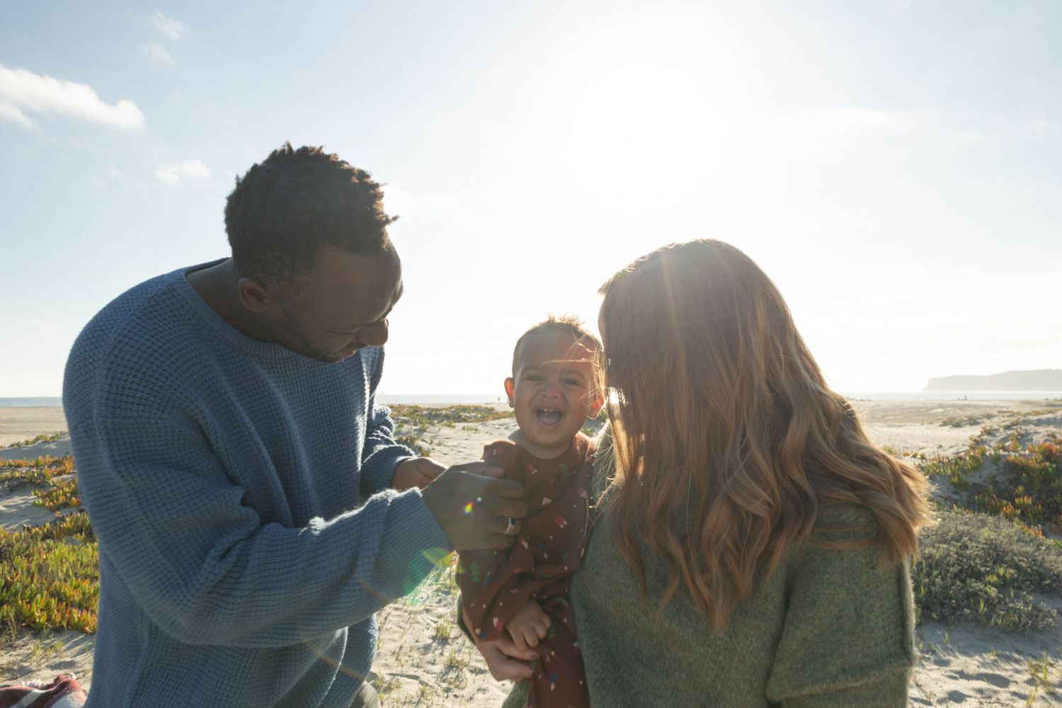 A family with a baby enjoys a sunny beach day together smiling and laughing together. Coronado beach family photographer.
