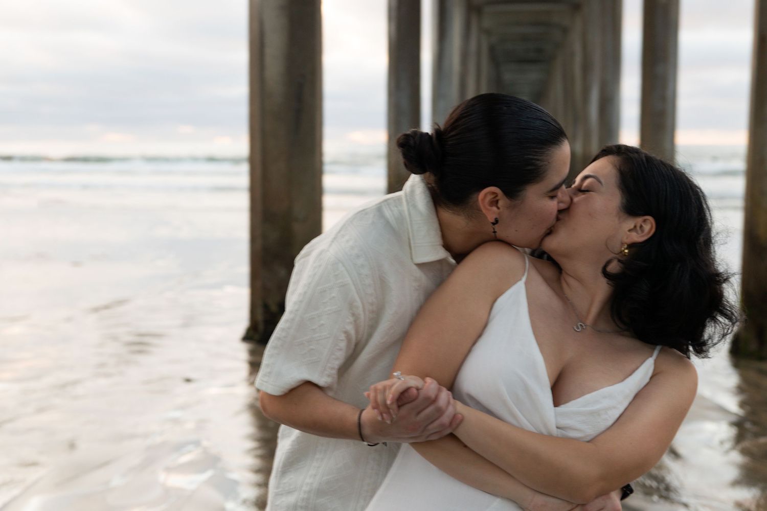 Two Fiancés, share a romantic kiss on the beach near a pier at sunset, one in white dress, one in white shirt. Engagement Photos.