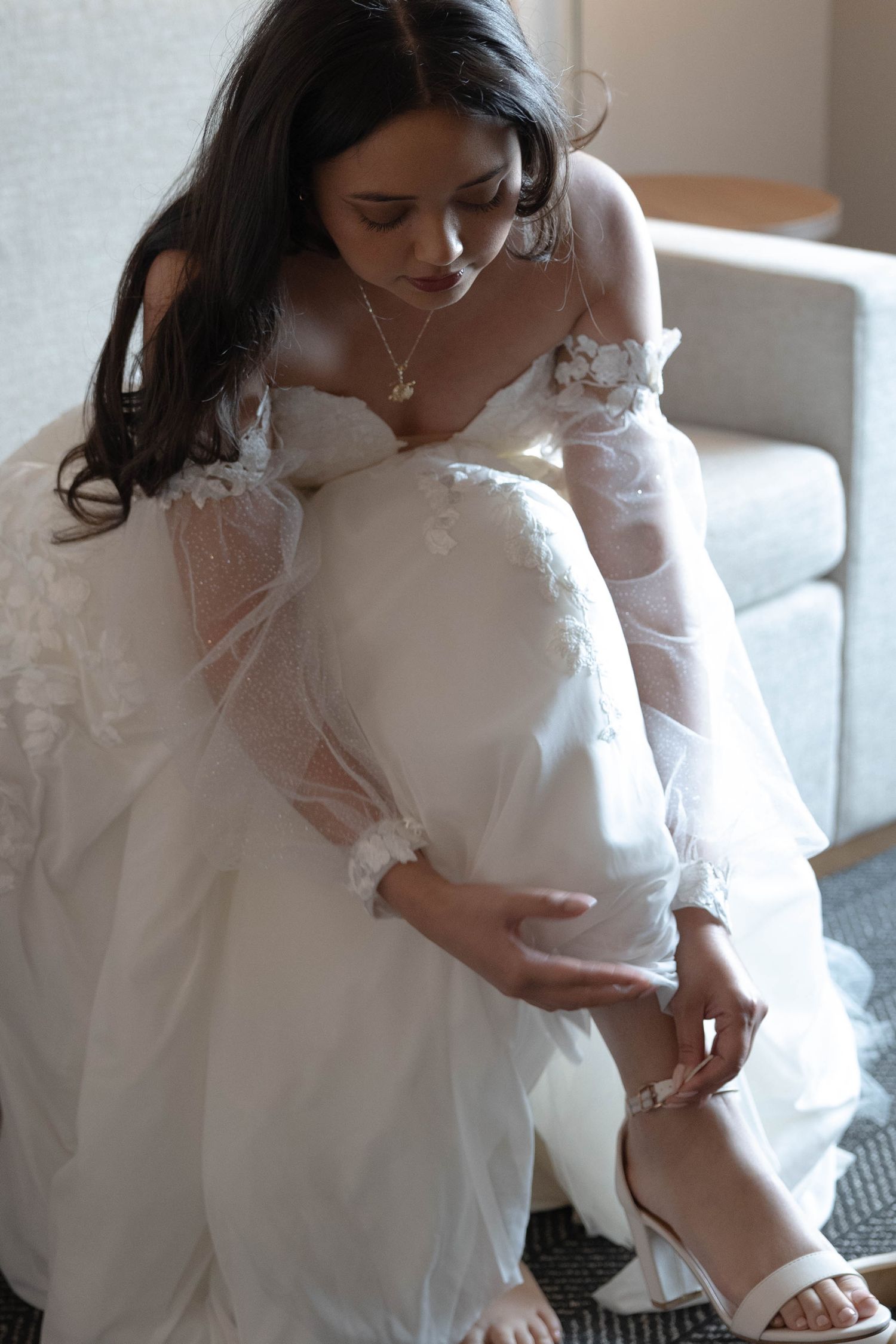 Bride in lace wedding gown fastening white heeled sandal while seated indoors.