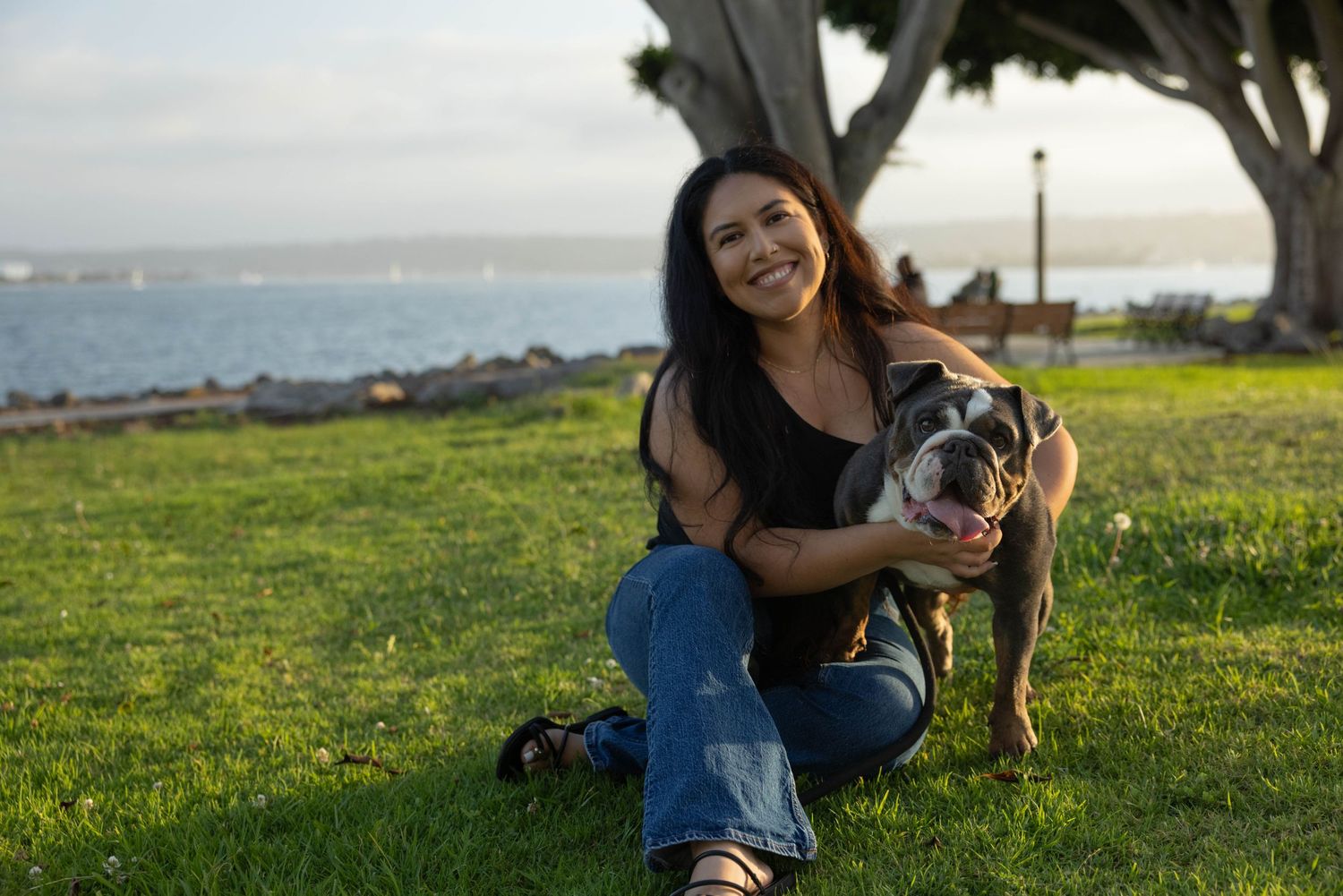Woman smiling with her bulldog on green grass near the ocean on a sunny day.