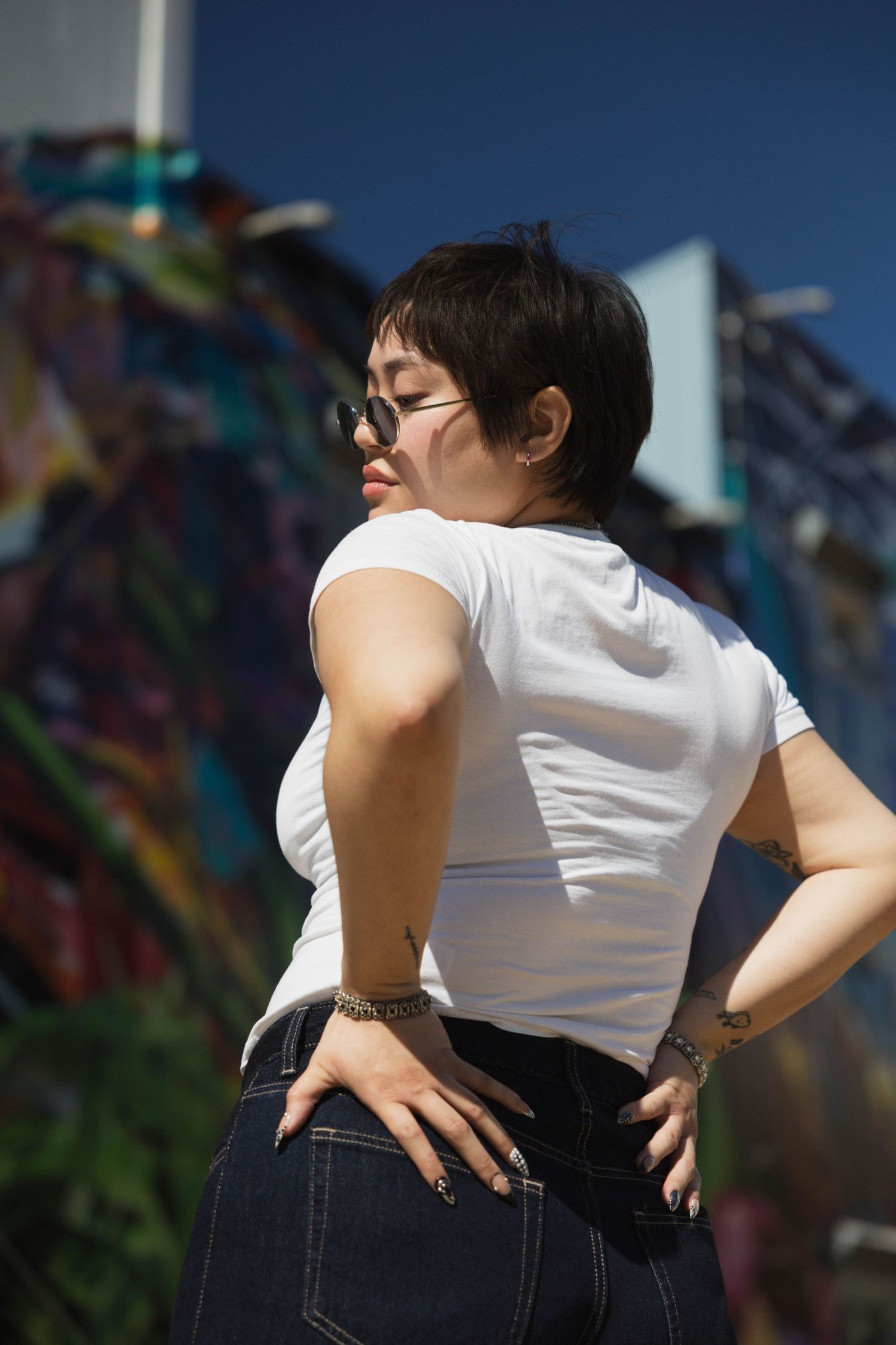 Stylish woman with short hair and sunglasses posing in white tee and jeans before colorful mural.