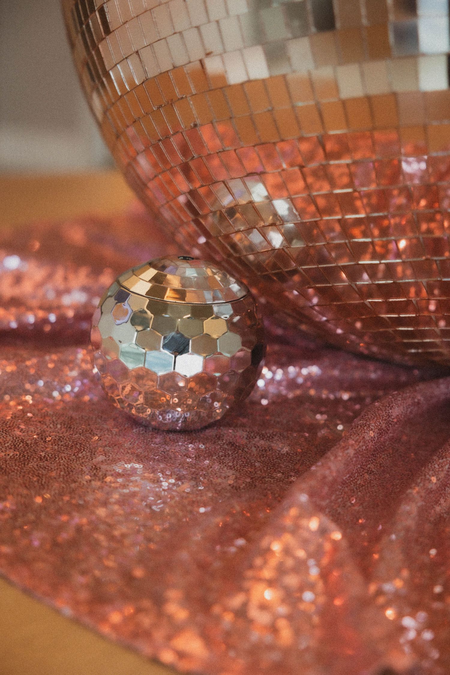 Close-up of a large rose gold disco ball beside a small mirrored sphere on a pink sequin fabric surface.