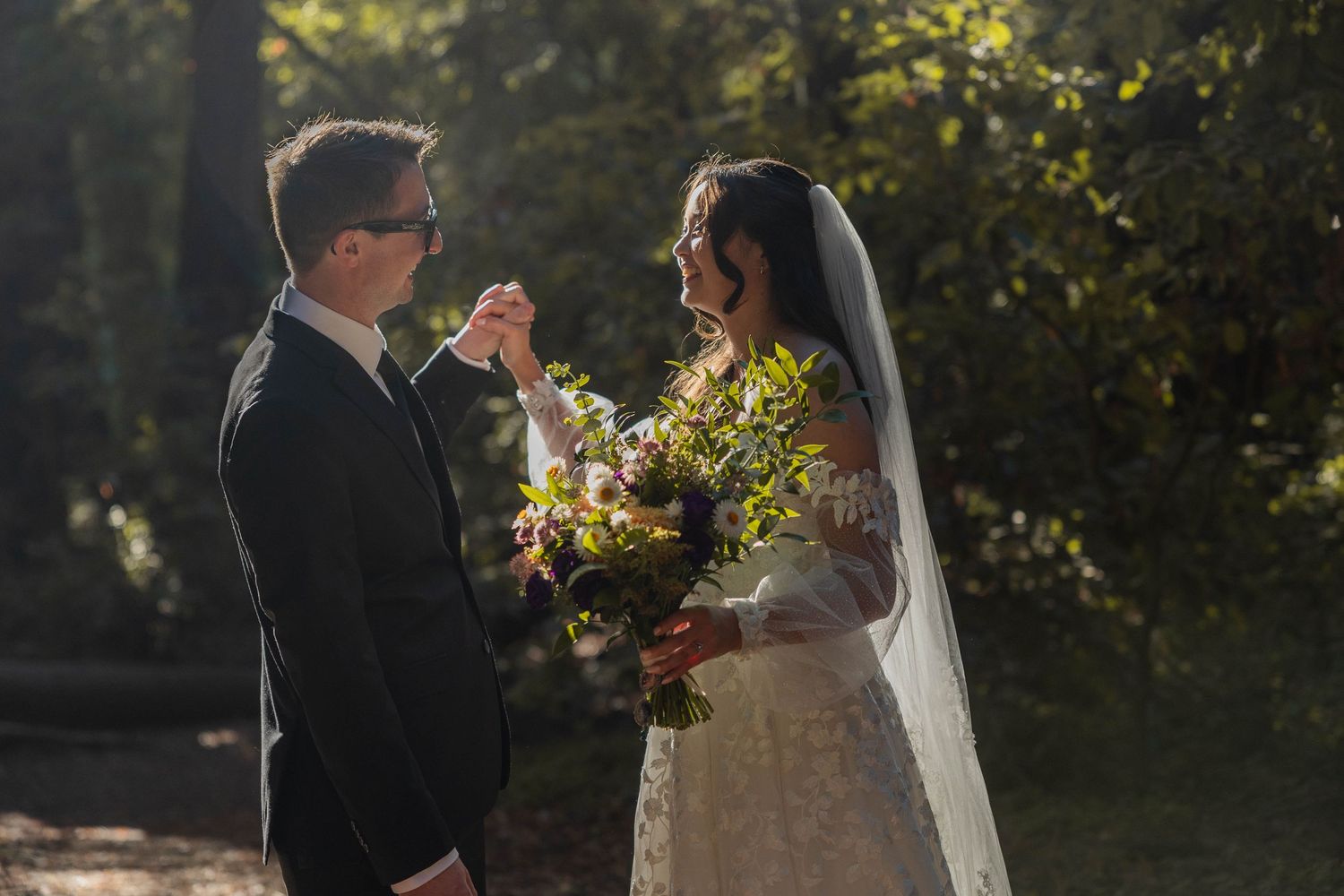 Bride and groom share a tender moment in a sunlit forest, bride holding a lush floral bouquet in a lace wedding gown.