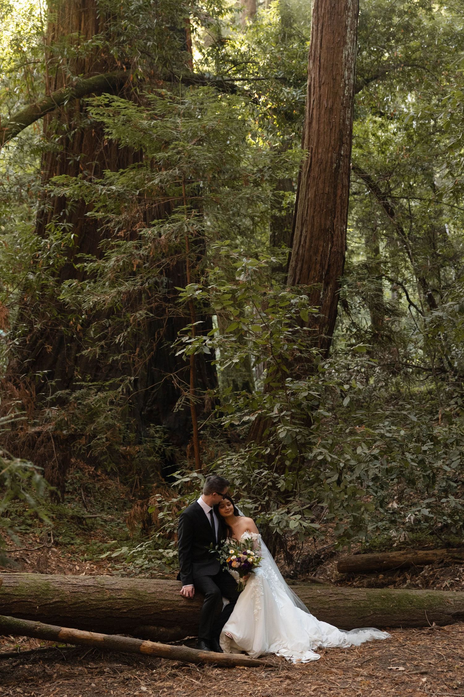 Bride and groom sit on a fallen log in a lush redwood forest, bride holding a bouquet in a white gown. Wedding photography.
