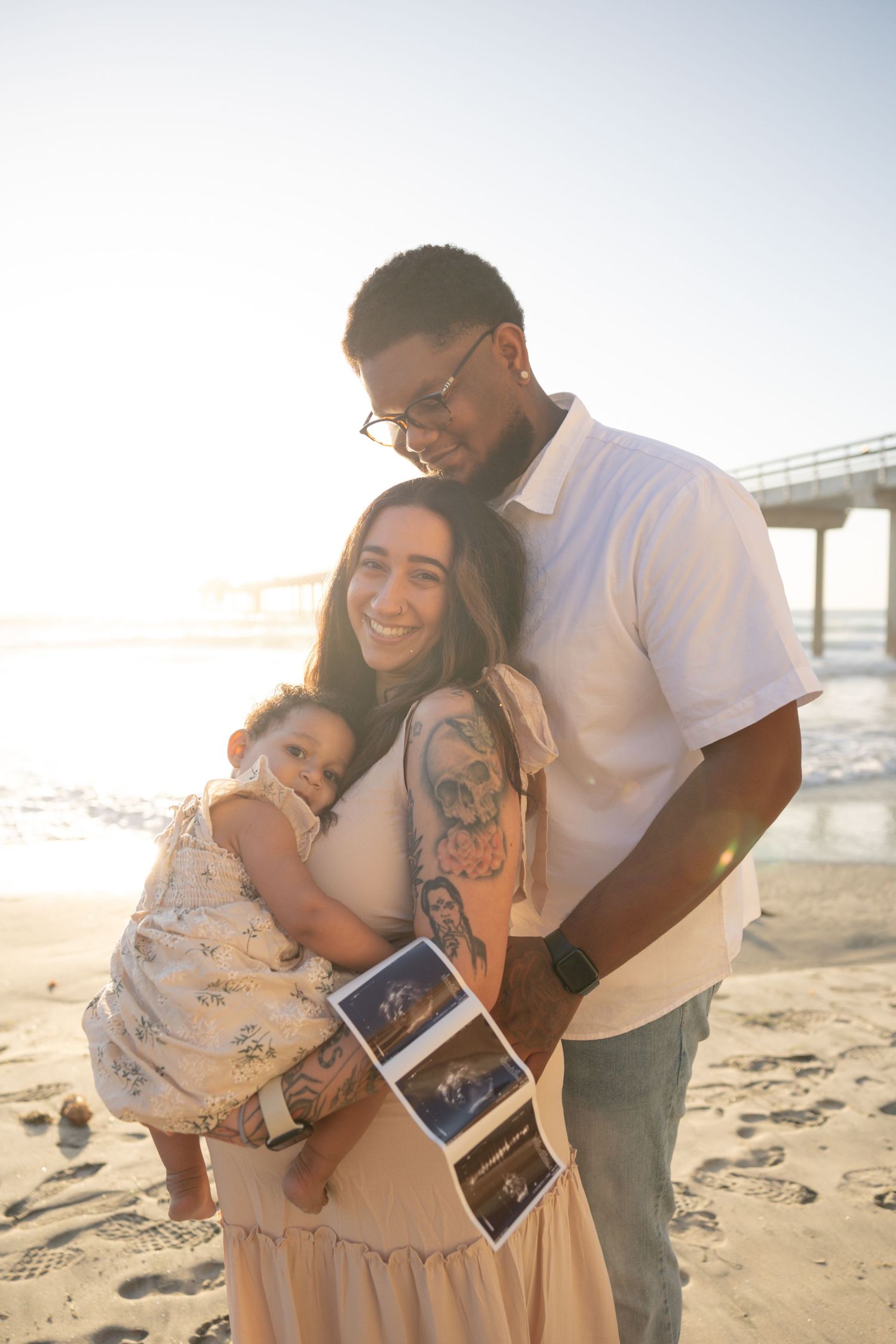 Happy couple with toddler on sunny beach holding ultrasound photos, announcing pregnancy near ocean pier. Maternity Photographer San Diego.