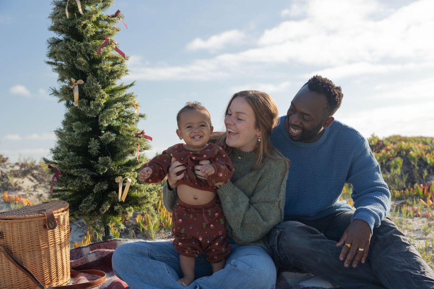 Happy interracial family with baby celebrating Christmas outdoors near decorated tree on sunny day.