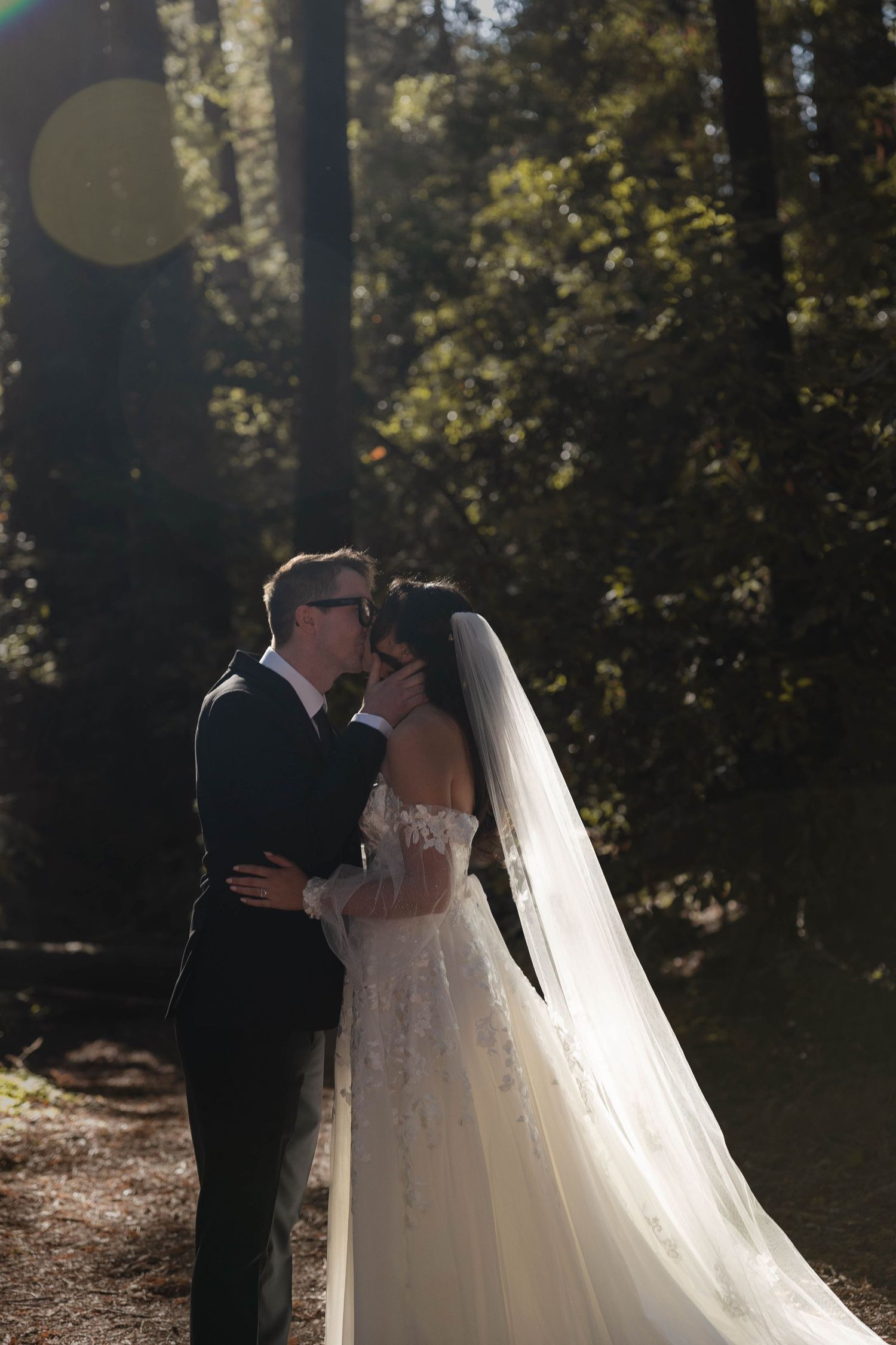 A bride and groom share a romantic kiss in a sunlit forest, her lace gown and long veil trailing behind them. Elopement Photographer.
