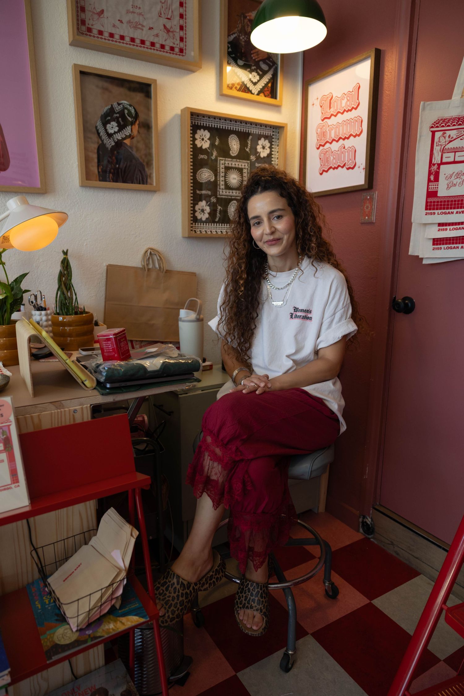 Young woman with curly hair sitting on stool in eclectic red and pink boutique shop surrounded by artwork.