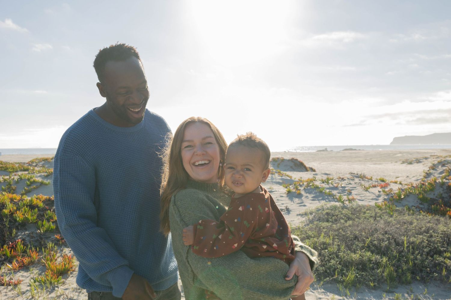 Family with baby smiling outdoors on a sunny beach with ocean views and natural coastal scenery. Coronado beach family photographer.