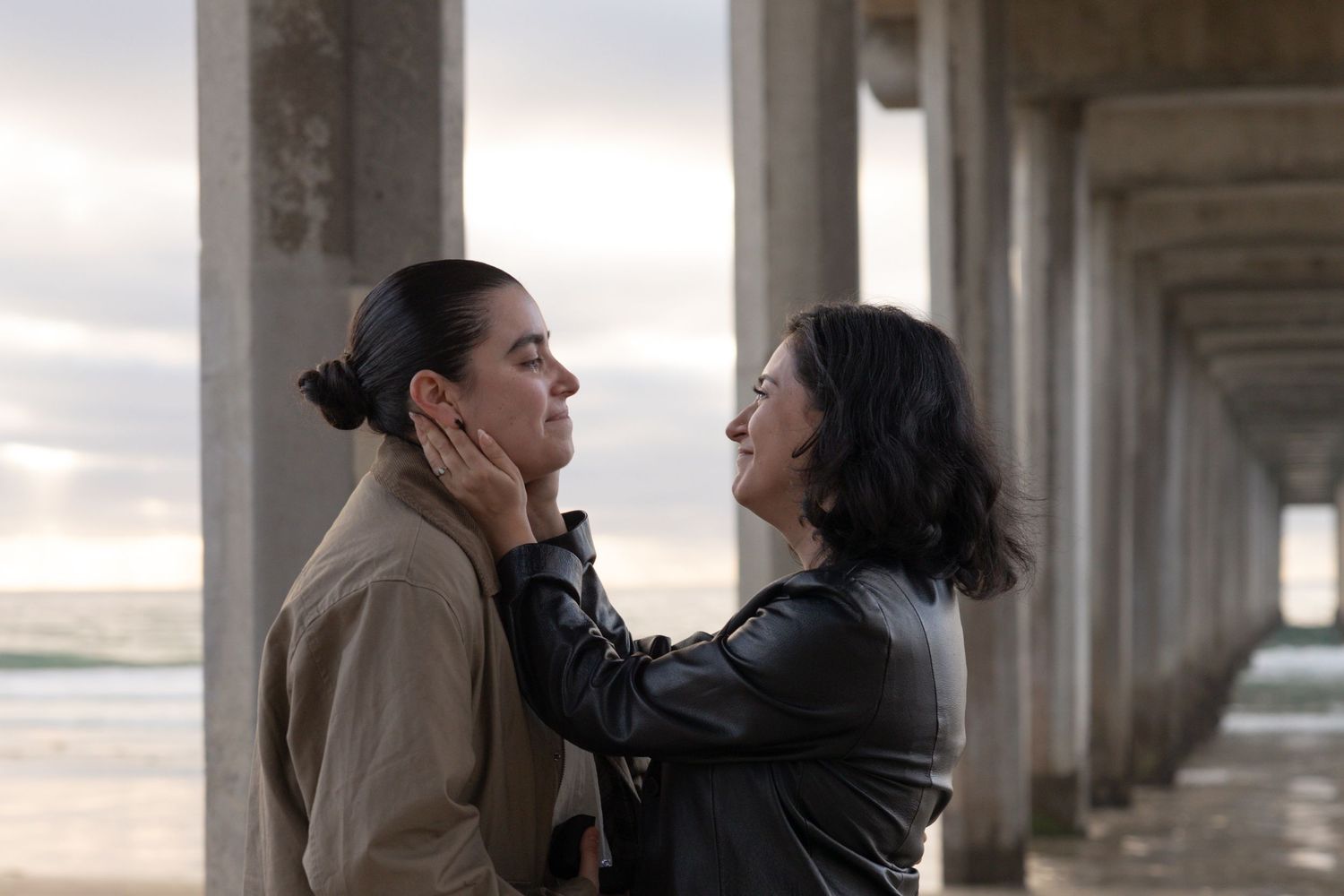 Two women share an intimate moment under a concrete pier by the ocean, one gently touching the other's face. La Jolla Shores, San Diego, CA.
