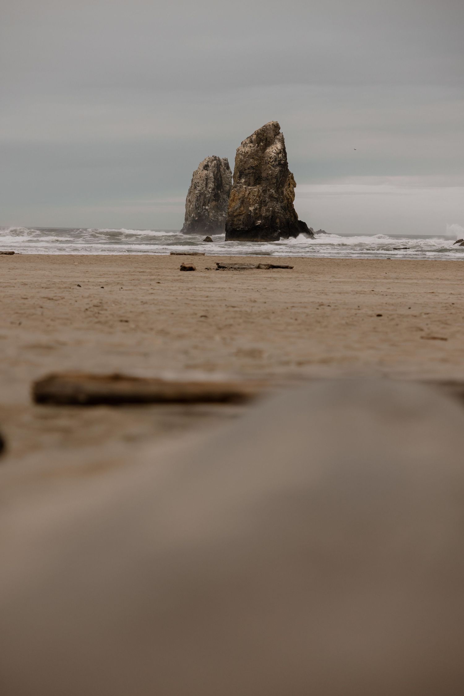 Two tall sea stacks rise from a sandy beach under overcast skies, with gentle waves and wet sand in the foreground.