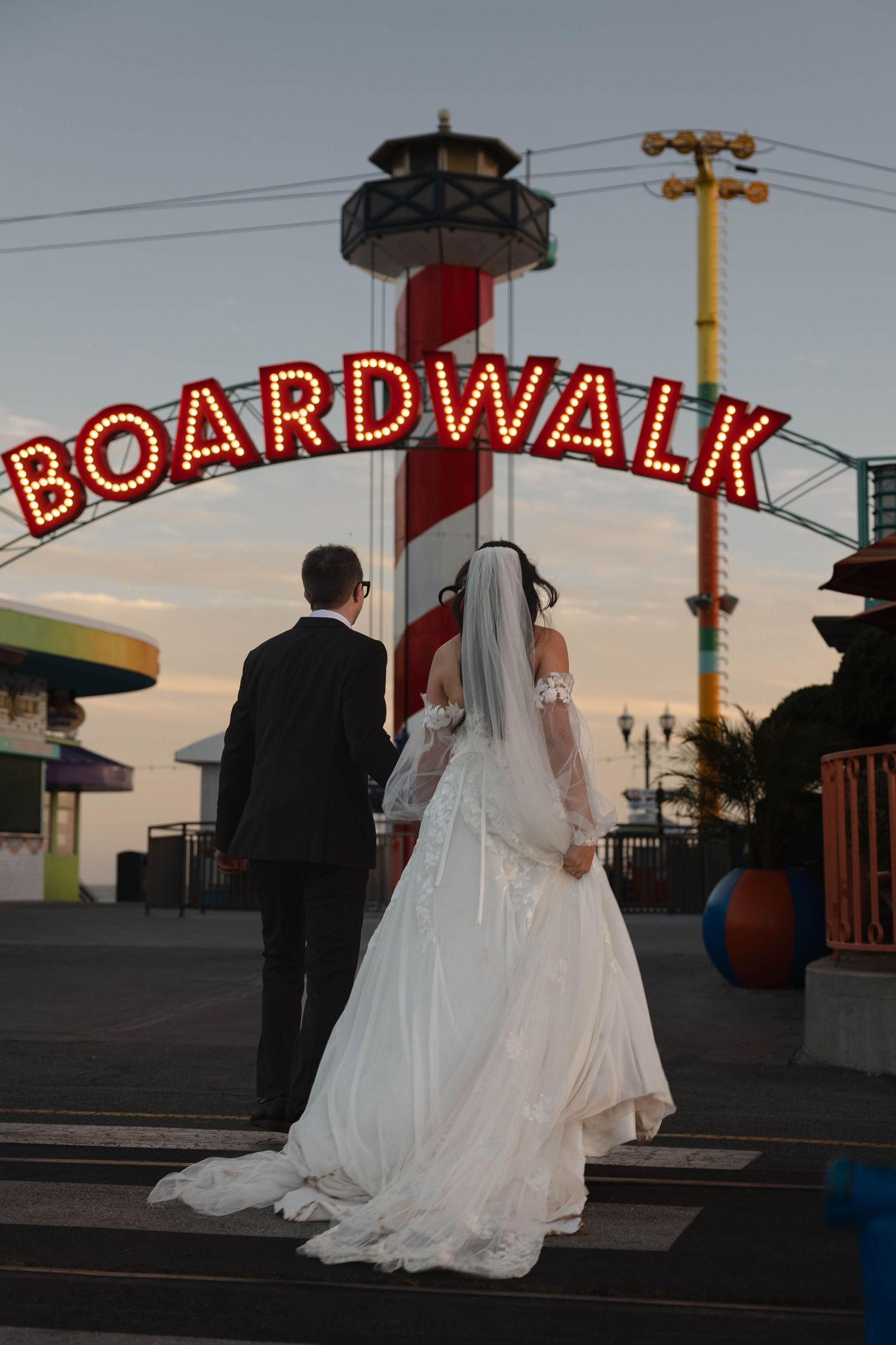 Bride and groom holding hands facing the illuminated Boardwalk sign at Santa Cruz amusement park at dusk.
