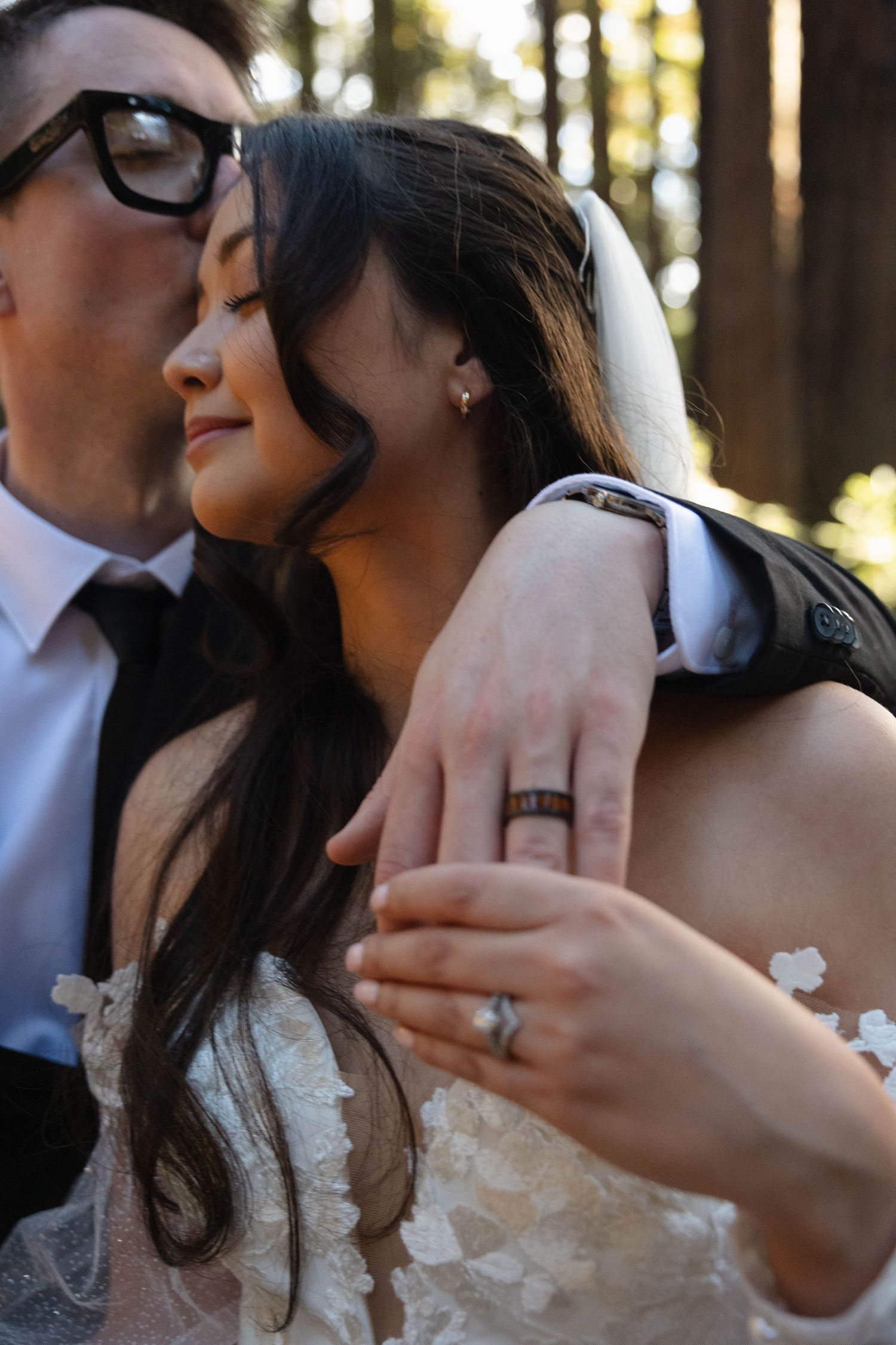 Close-up of bride and groom embracing, showcasing wedding rings, bride in lace gown with groom in black suit.