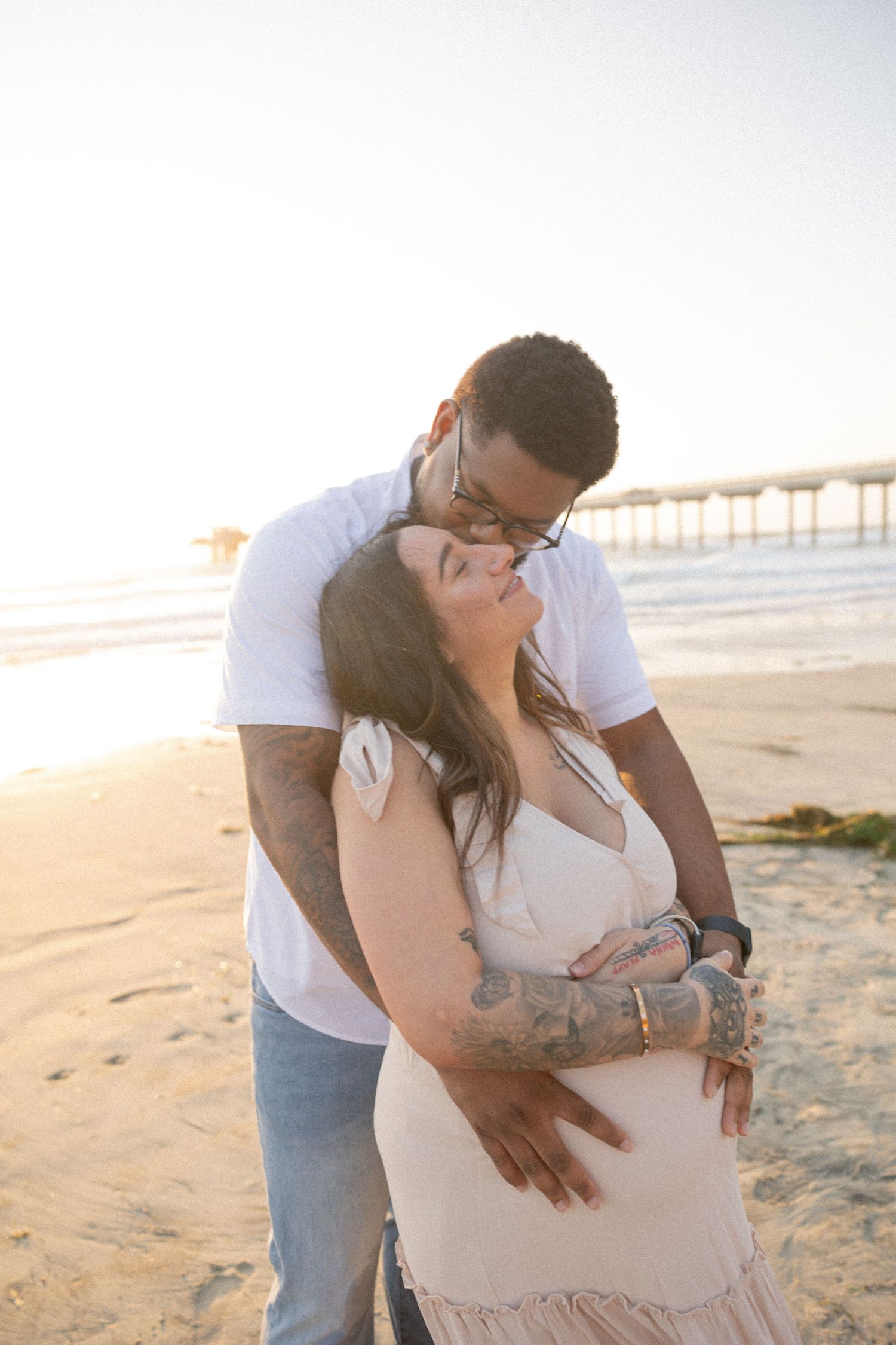 Couple embracing on a sunny beach near a pier, man kissing woman's cheek, golden hour. Maternity Photographer San Diego California.