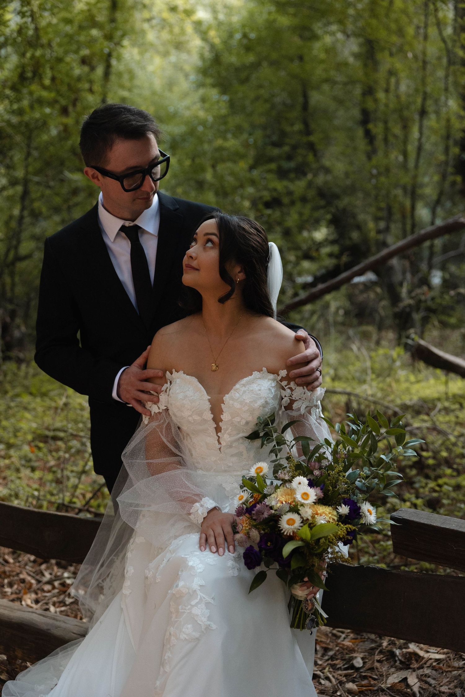 Groom in black suit embraces bride in lace gown holding wildflower bouquet in a forest setting. Wedding photography.