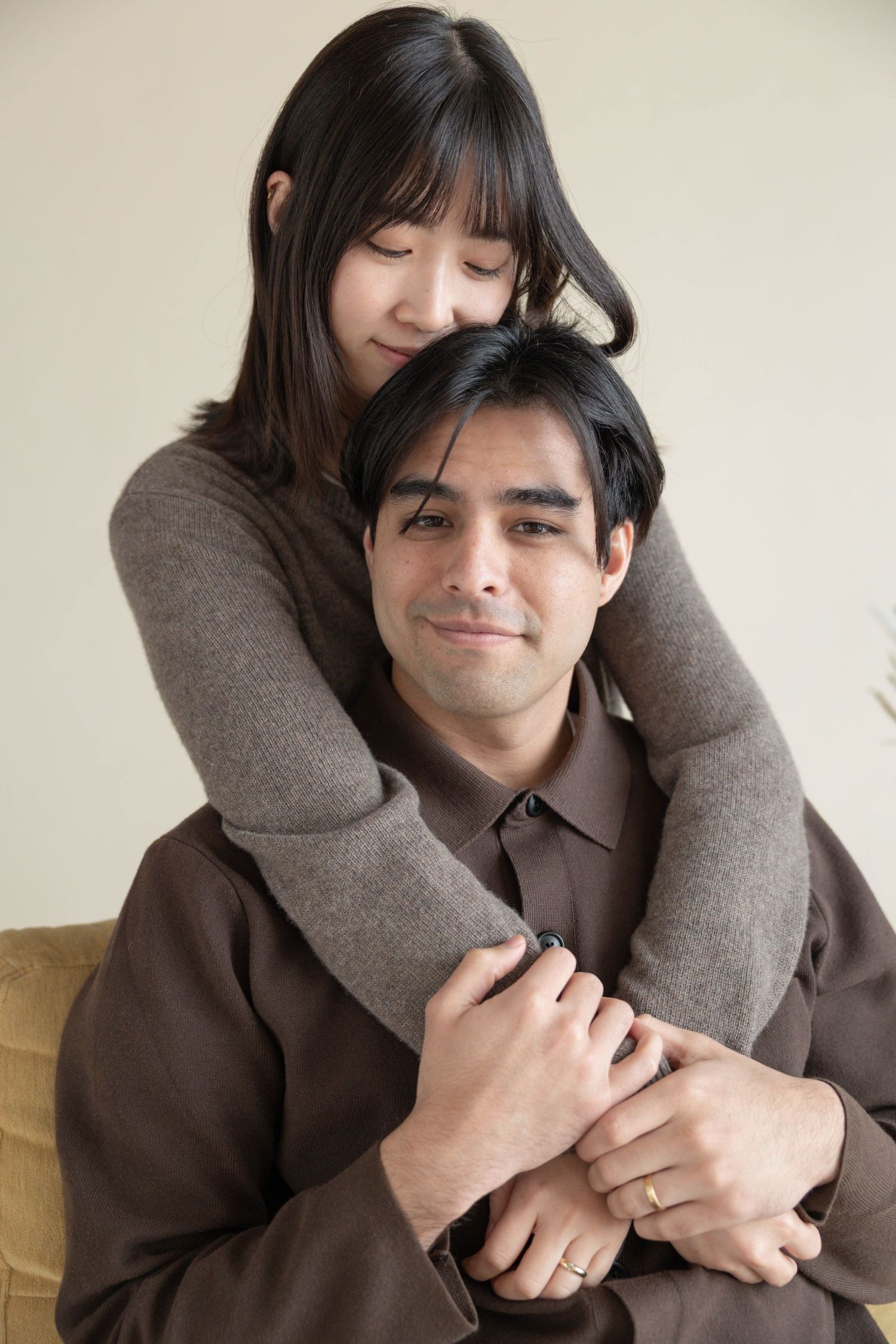 A woman embraces a man from behind on a sofa, both wearing brown clothing in a warm, affectionate pose. Couples Portraits San Diego Ca