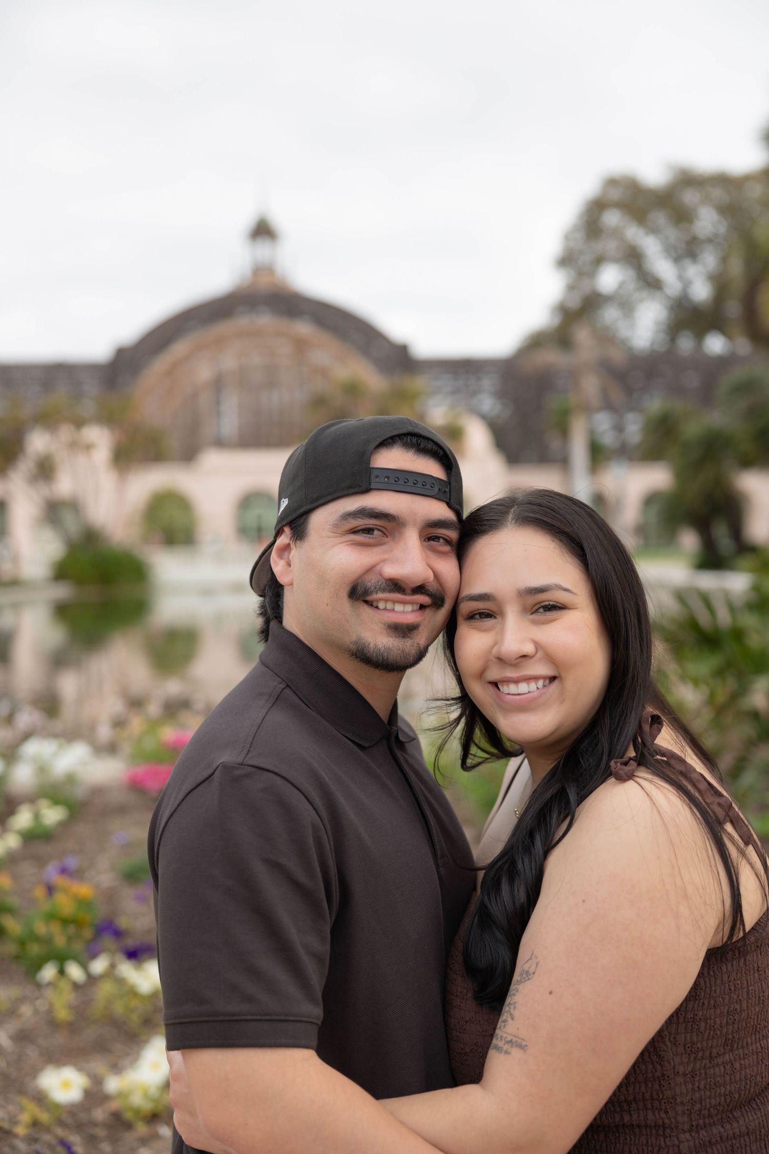 Smiling couple embraces in front of the Balboa Park Botanical Building with a lily pond in background.