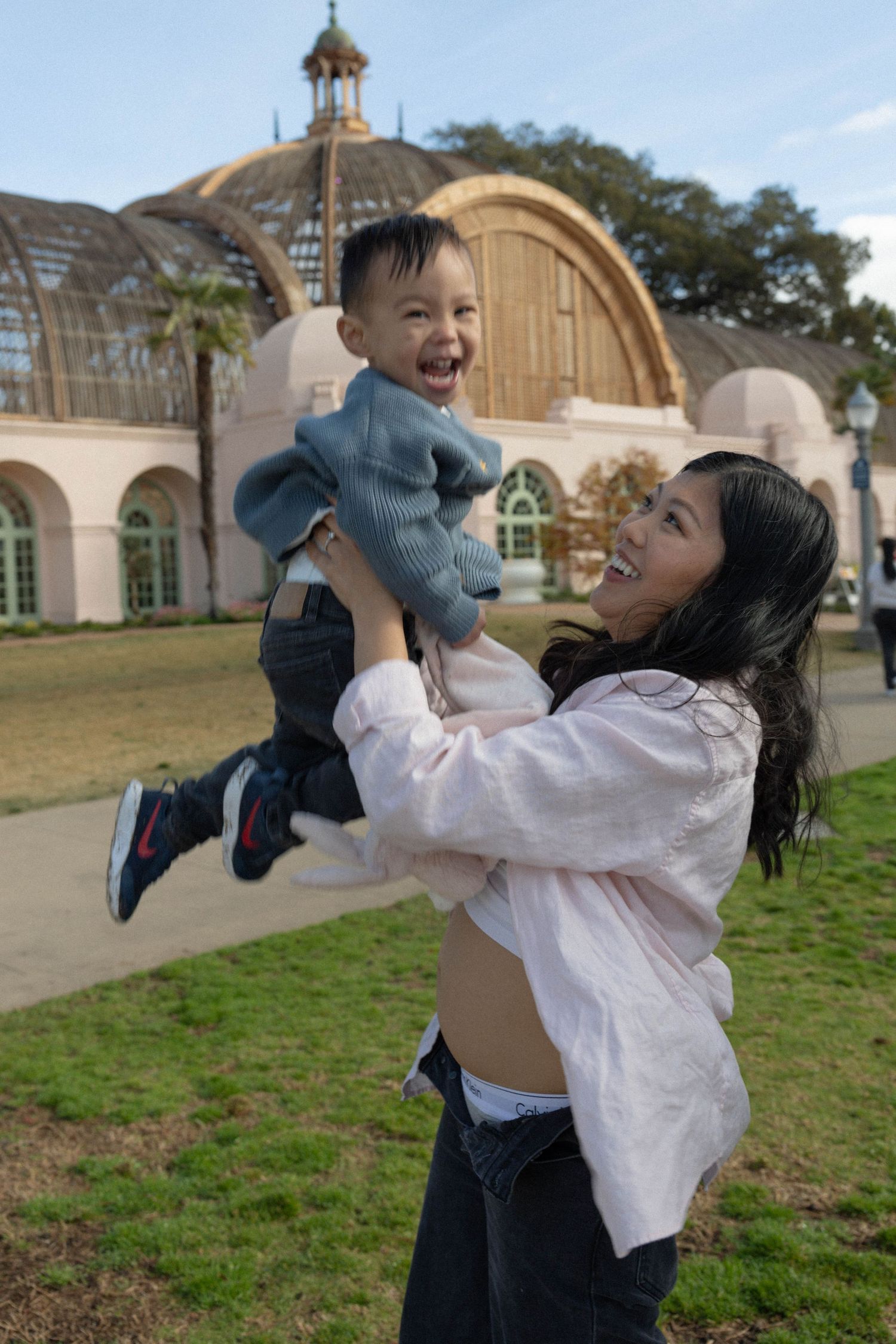 A joyful mother lifts her laughing toddler boy outdoors near Balboa Botanical Building. Family photographer San Diego California.