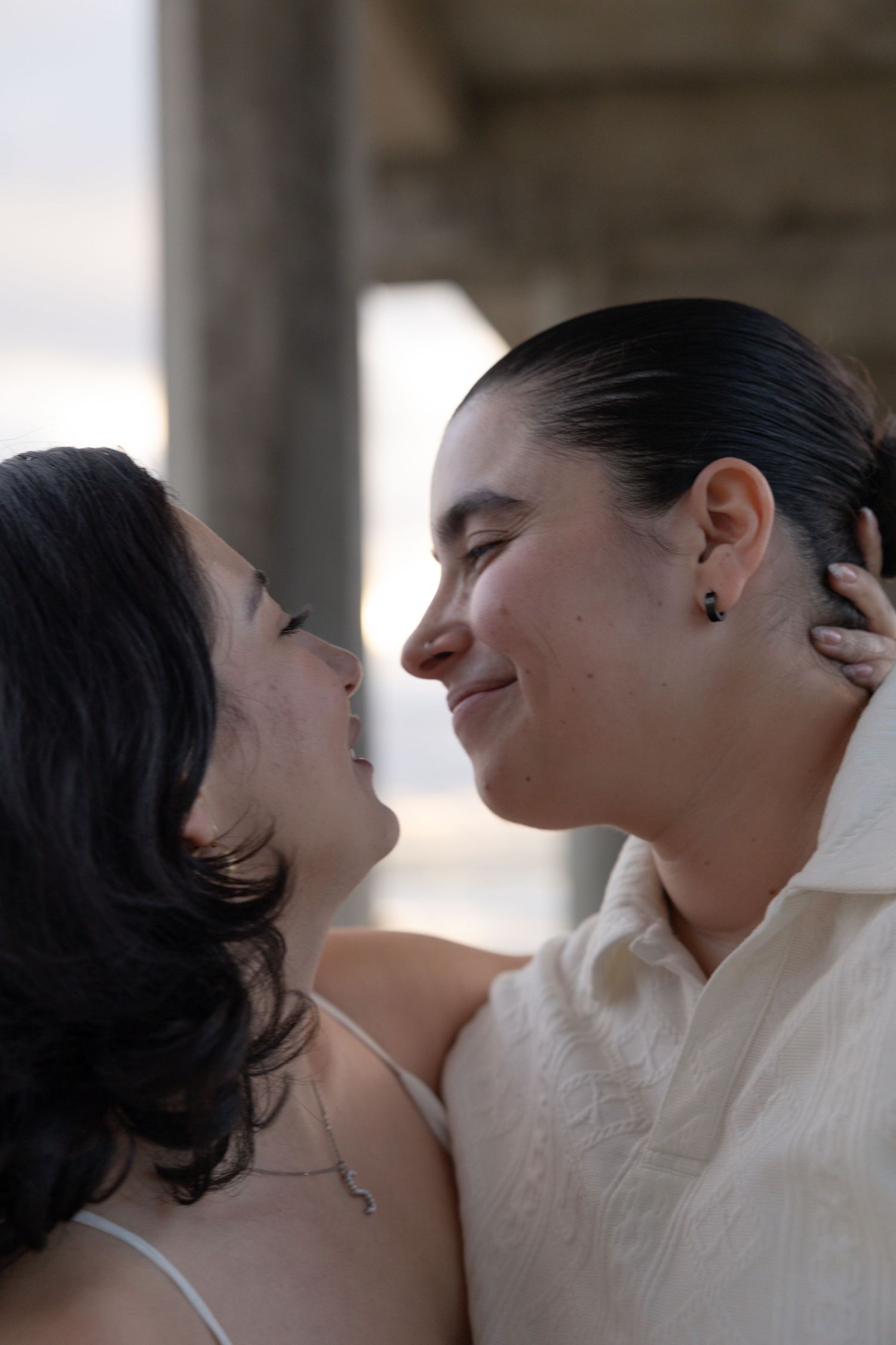 Two women share a romantic moment, smiling and about to kiss, dressed in white attire at La Jolla Shores. San Diego Engagement Photographer.