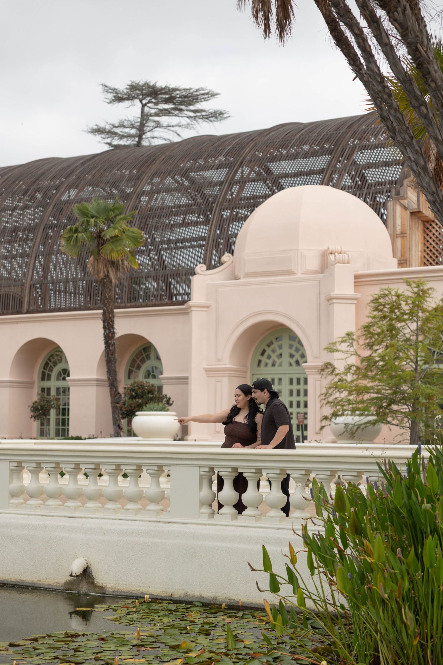 Couple poses romantically by a lily pond in front of a pink arched building with a large greenhouse dome.