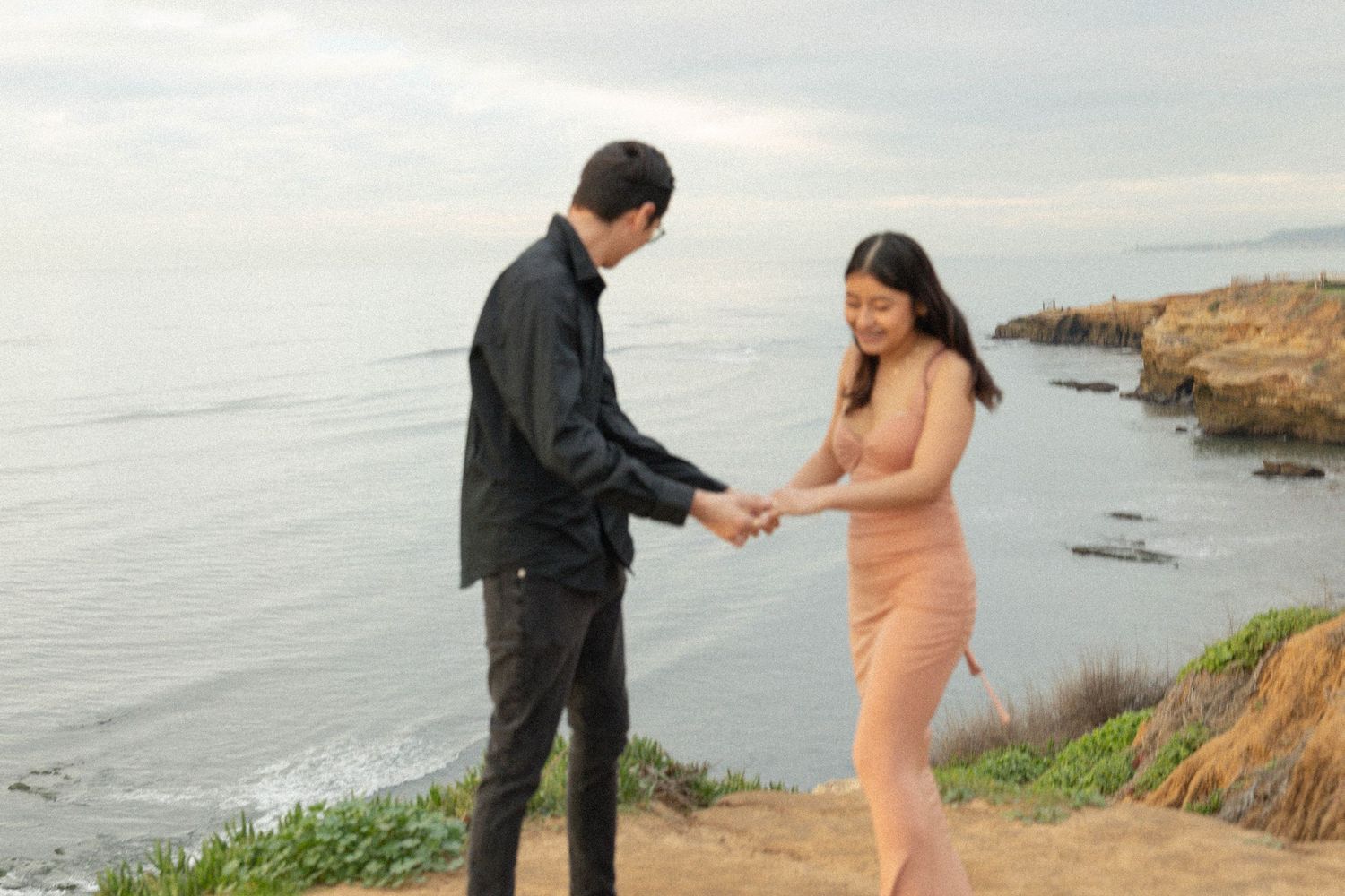 Couple holding hands on a coastal cliff overlooking the ocean on a cloudy day. San Diego Engagement Photographer.
