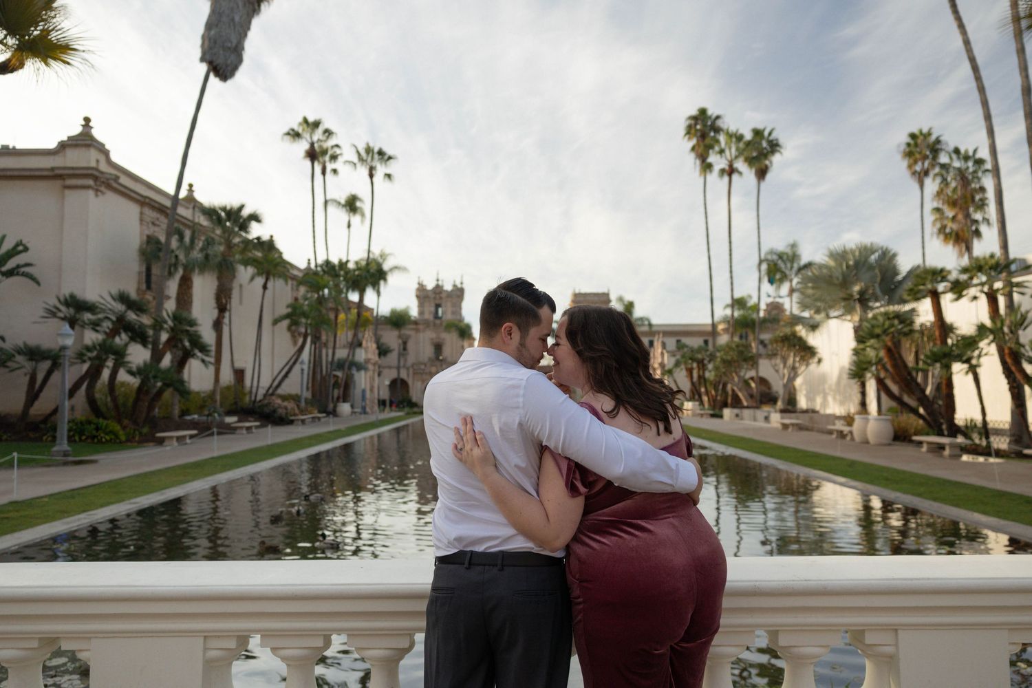 Couple embracing romantically at Balboa Park reflecting pool, surrounded by palm trees, in elegant engagement photo. Couples photography.