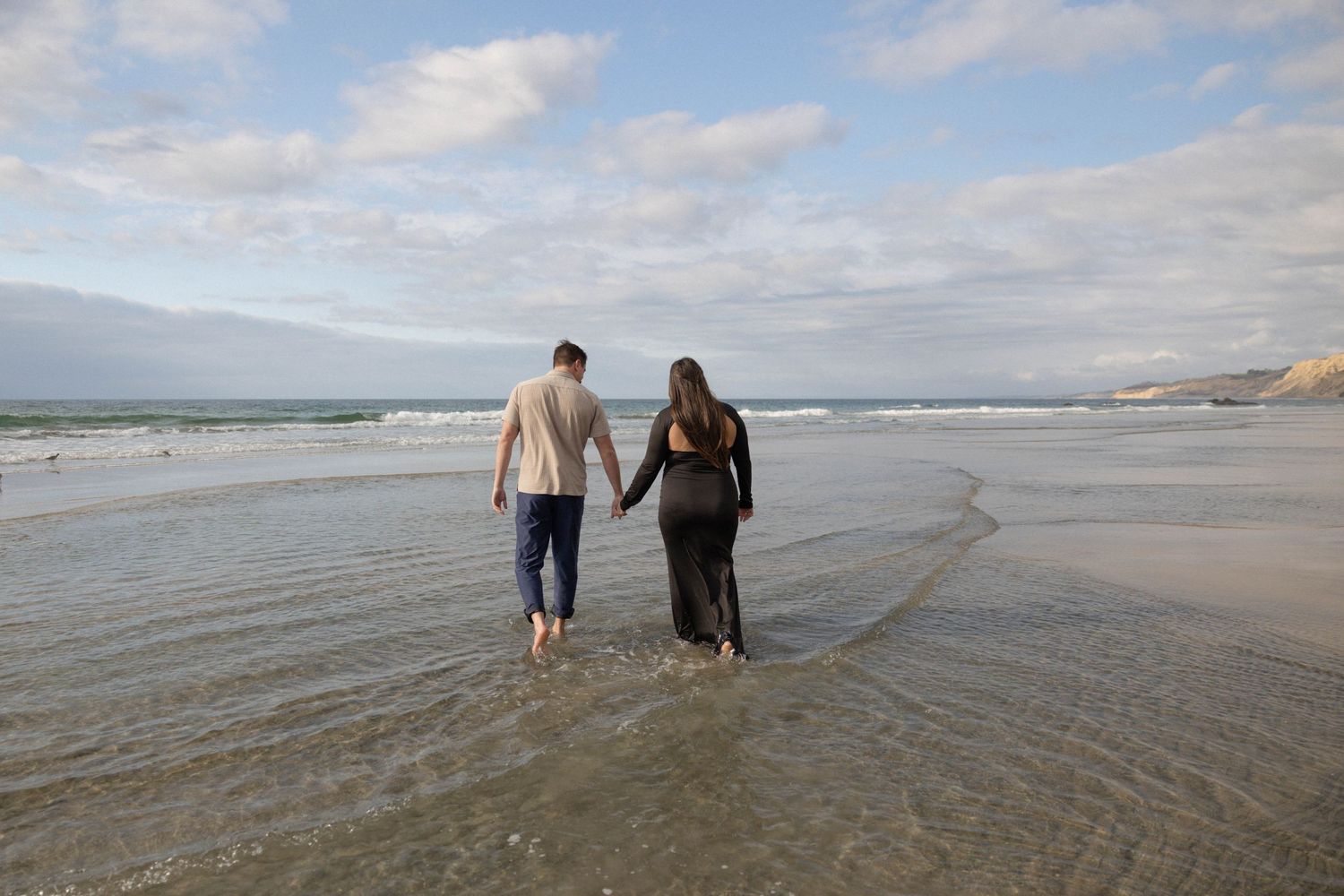 Couple holding hands walking barefoot along a sandy beach with ocean waves and cloudy sky backdrop. La Jolla San Diego Photographer.