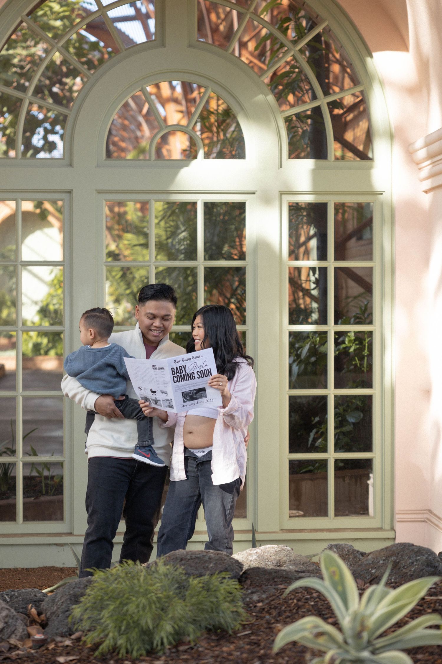 Family with toddler reads baby announcement newspaper outside elegant building with arched windows. Maternity Photographer San Diego.