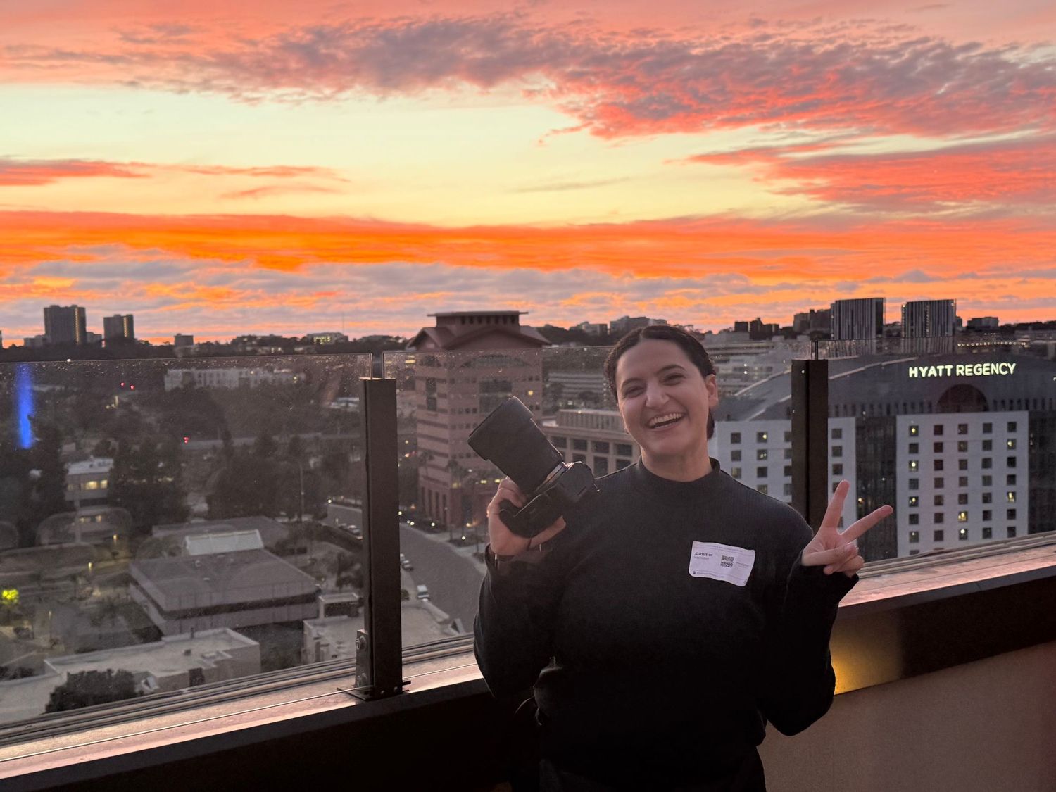 Woman smiling at sunset on a rooftop balcony near Hyatt Regency, holding a camera with a vibrant orange sky.