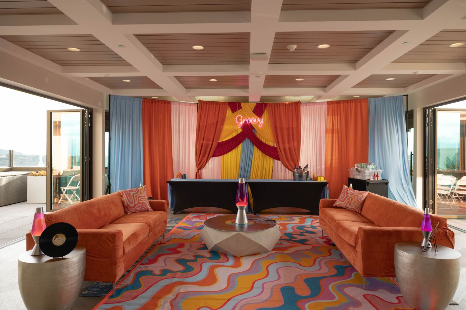 Vibrant retro lounge with orange velvet sofas, psychedelic wavy rug, and colorful draped stage backdrop with neon sign.