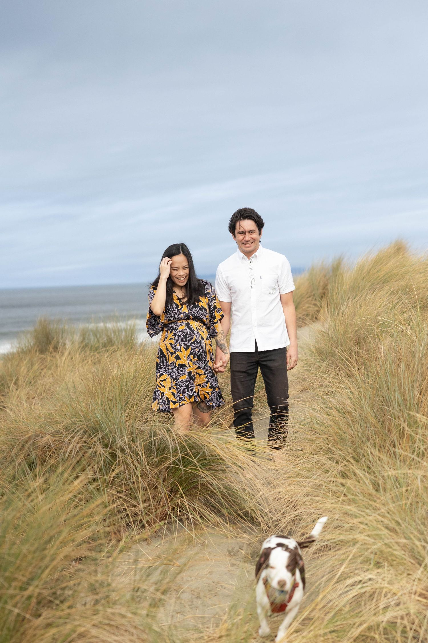 Couple walking with their dog through coastal sand dunes, woman in floral dress and man in white shirt, beach backdrop. Family Photographer.