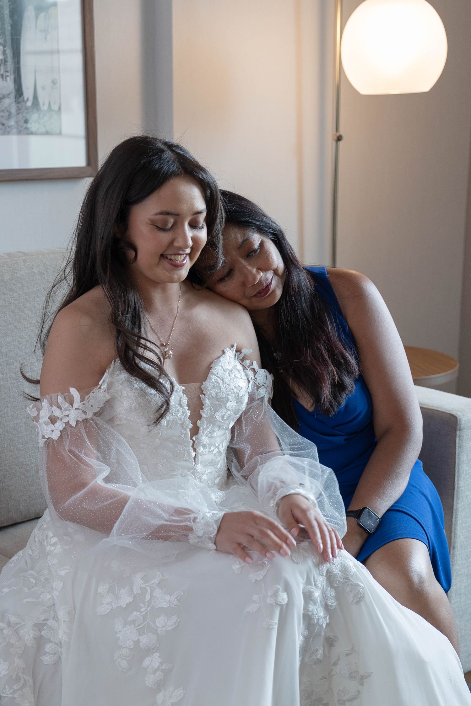 Bride in floral lace gown shares tender moment with bridesmaid in blue dress.