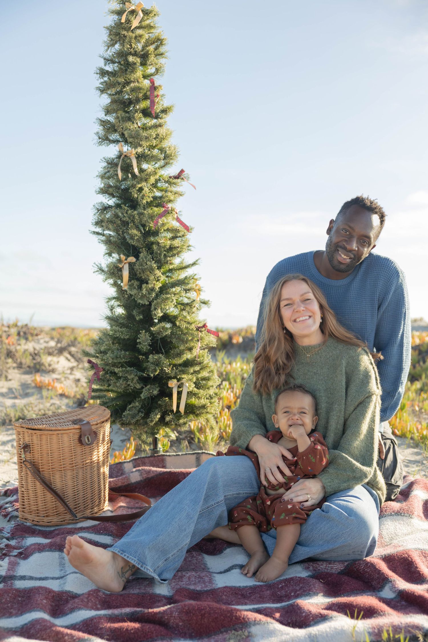 Interracial family with baby sitting on blanket by decorated Christmas tree outdoors on sunny day.