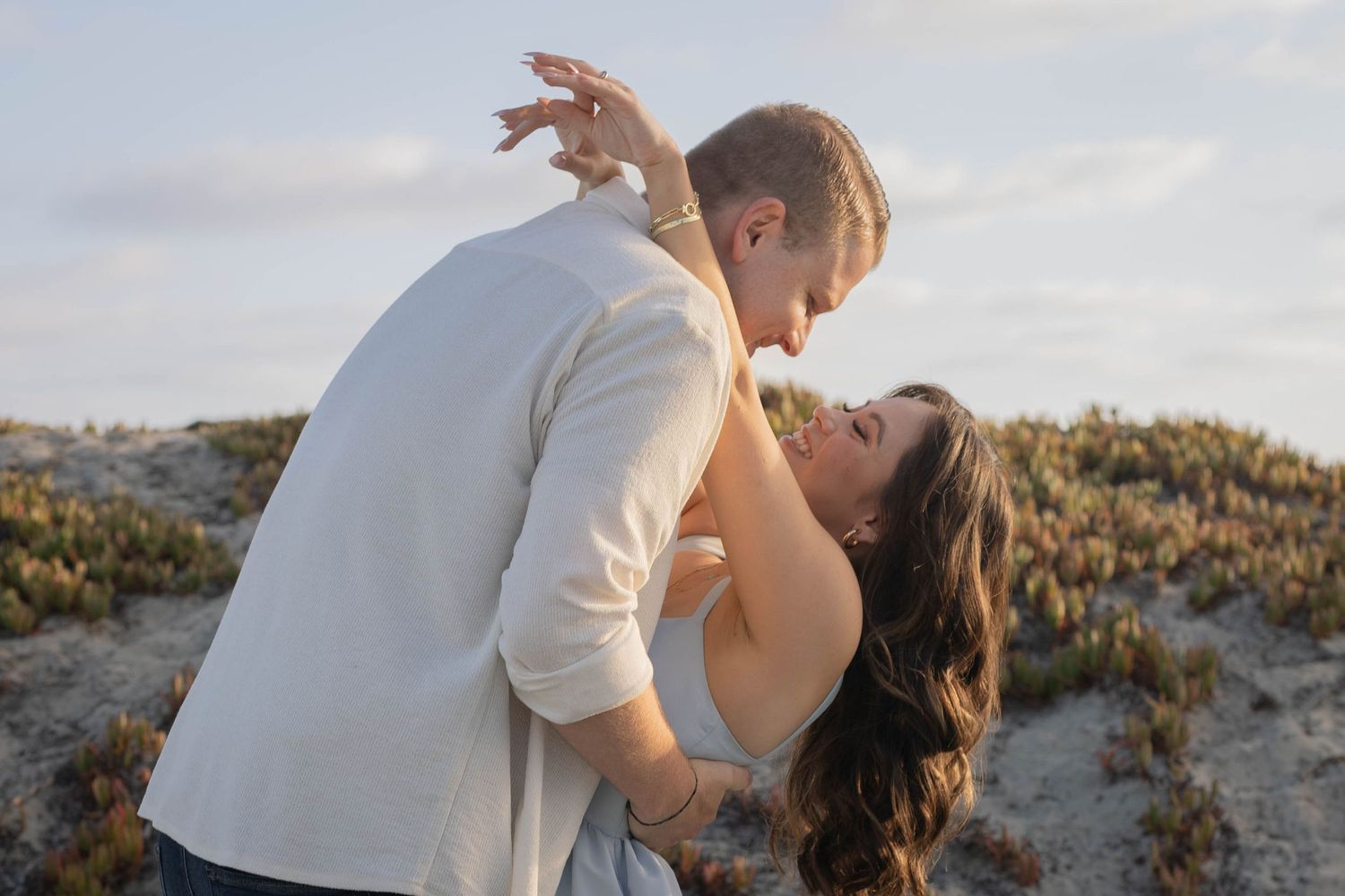 Couple dancing romantically on sandy beach dunes at golden hour, woman laughing joyfully as man dips her gracefully.