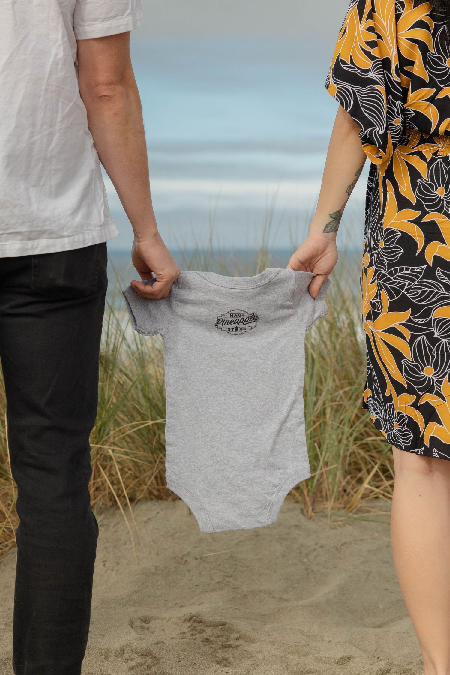 Couple holding a gray baby onesie on a sandy beach, pregnancy announcement photo with ocean backdrop. Family Photographer.