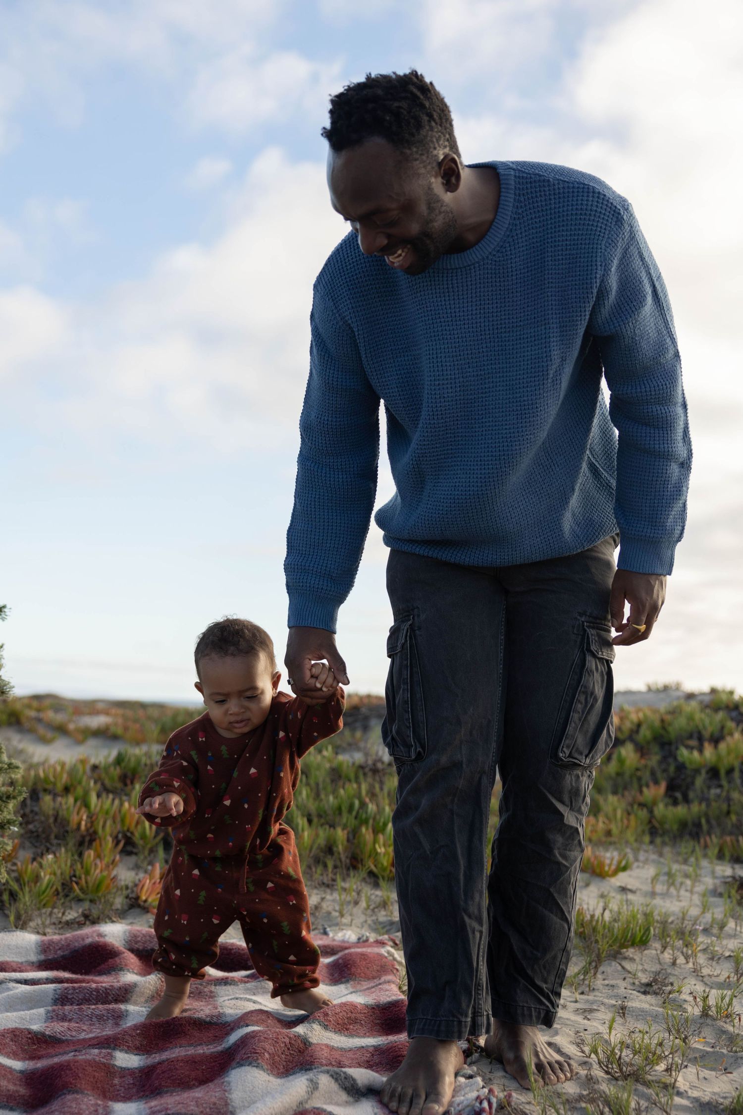 A smiling father helps his toddler walk on a blanket at the beach, both enjoying a sunny outdoor day. Family Photographer.