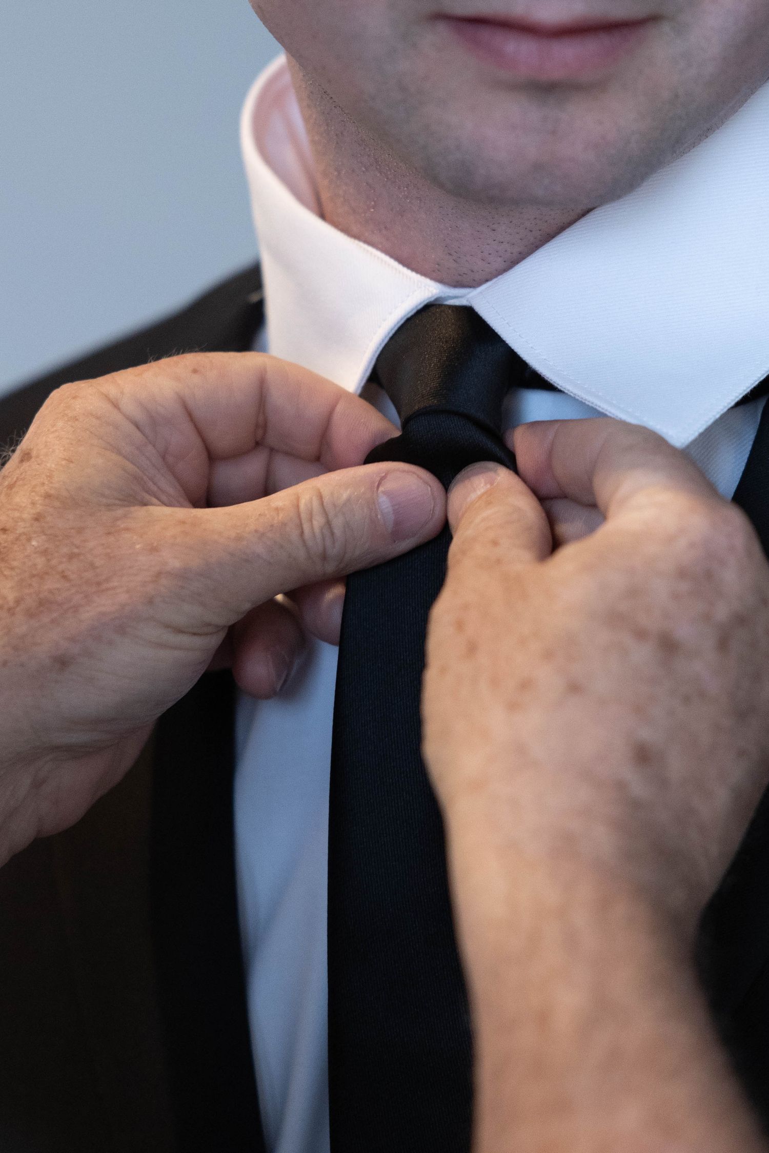 Hands adjusting black necktie on groom in white dress shirt and black suit.