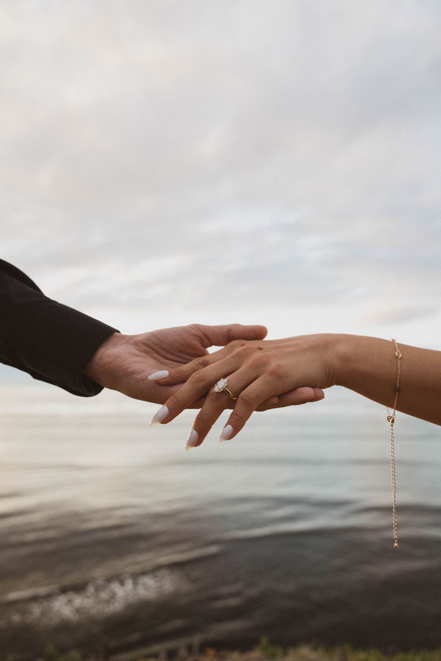 Couple holding hands near ocean, bride wearing diamond ring and gold bracelet.