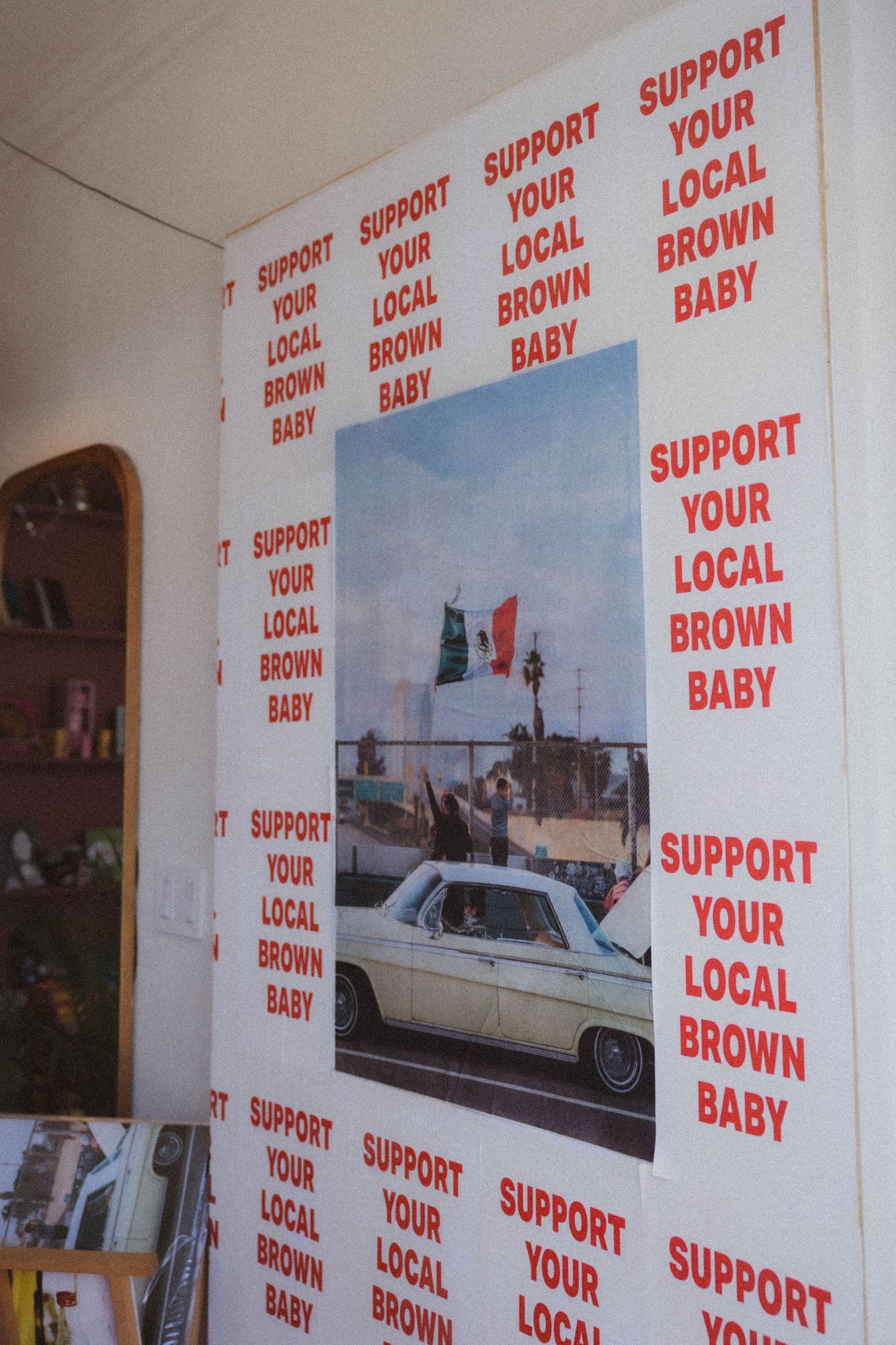Wall decorated with repeating red and white Support Your Local Brown Baby posters surrounding a Mexican flag photograph.