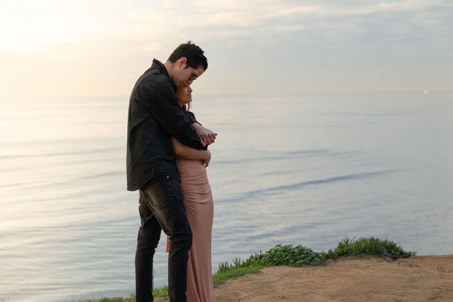 Couple sharing a tender embrace on a cliff overlooking a calm ocean at sunset.