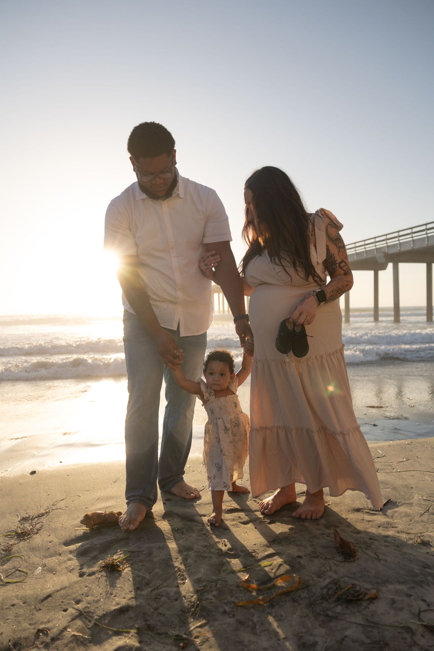 Family of three holding hands on sandy beach at sunset near pier with tiny baby shoes. Sunset. Maternity Photographer San Diego California.