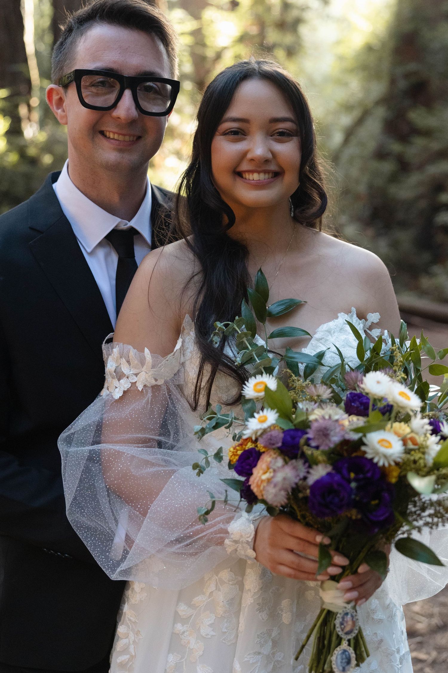 A smiling bride holding a colorful bouquet of wildflowers stands with a groom in a black suit and glasses outdoors. Wedding photography.