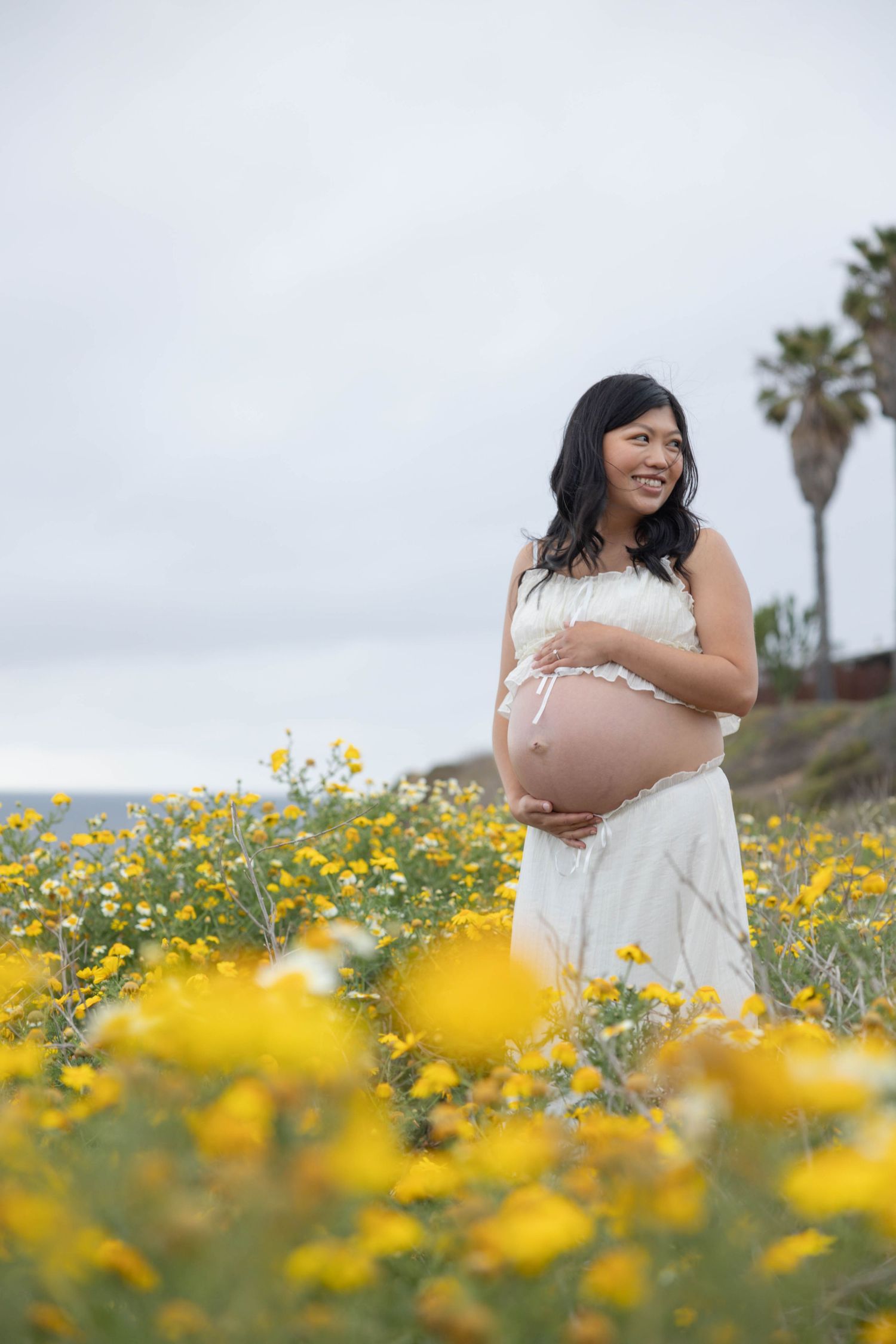 Pregnant woman in white dress holds her baby bump while standing in a field of yellow wildflowers.