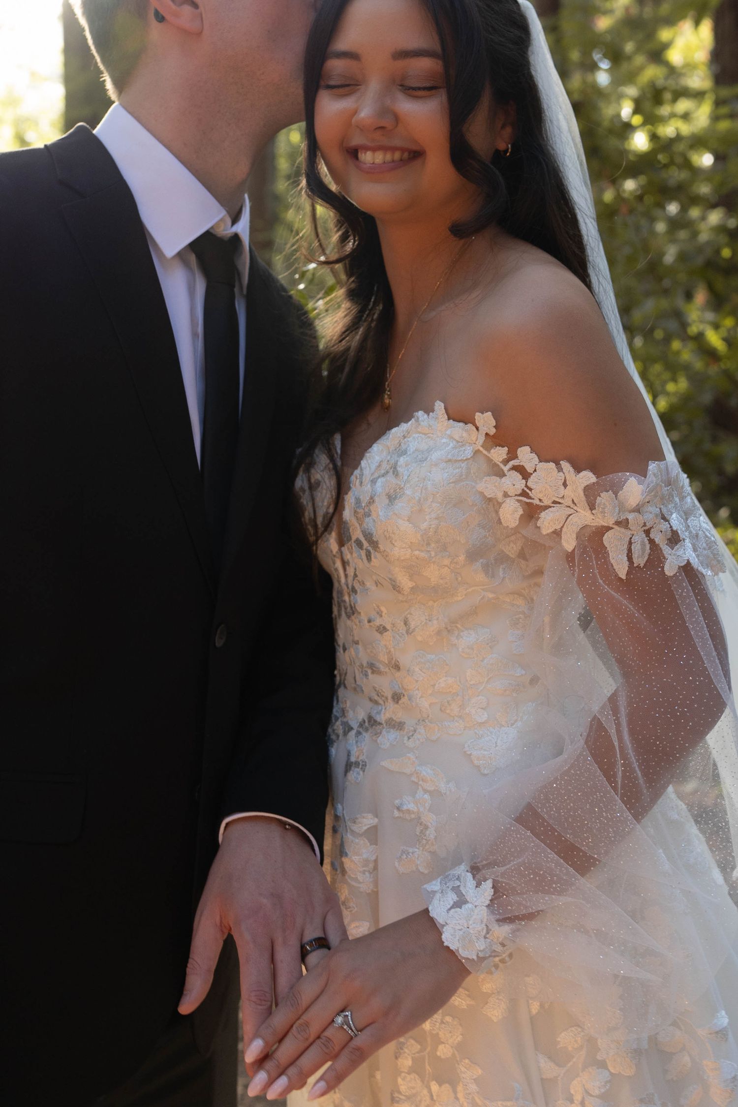 Close-up of smiling bride in floral lace off-shoulder gown with veil, groom kissing her cheek outdoors.