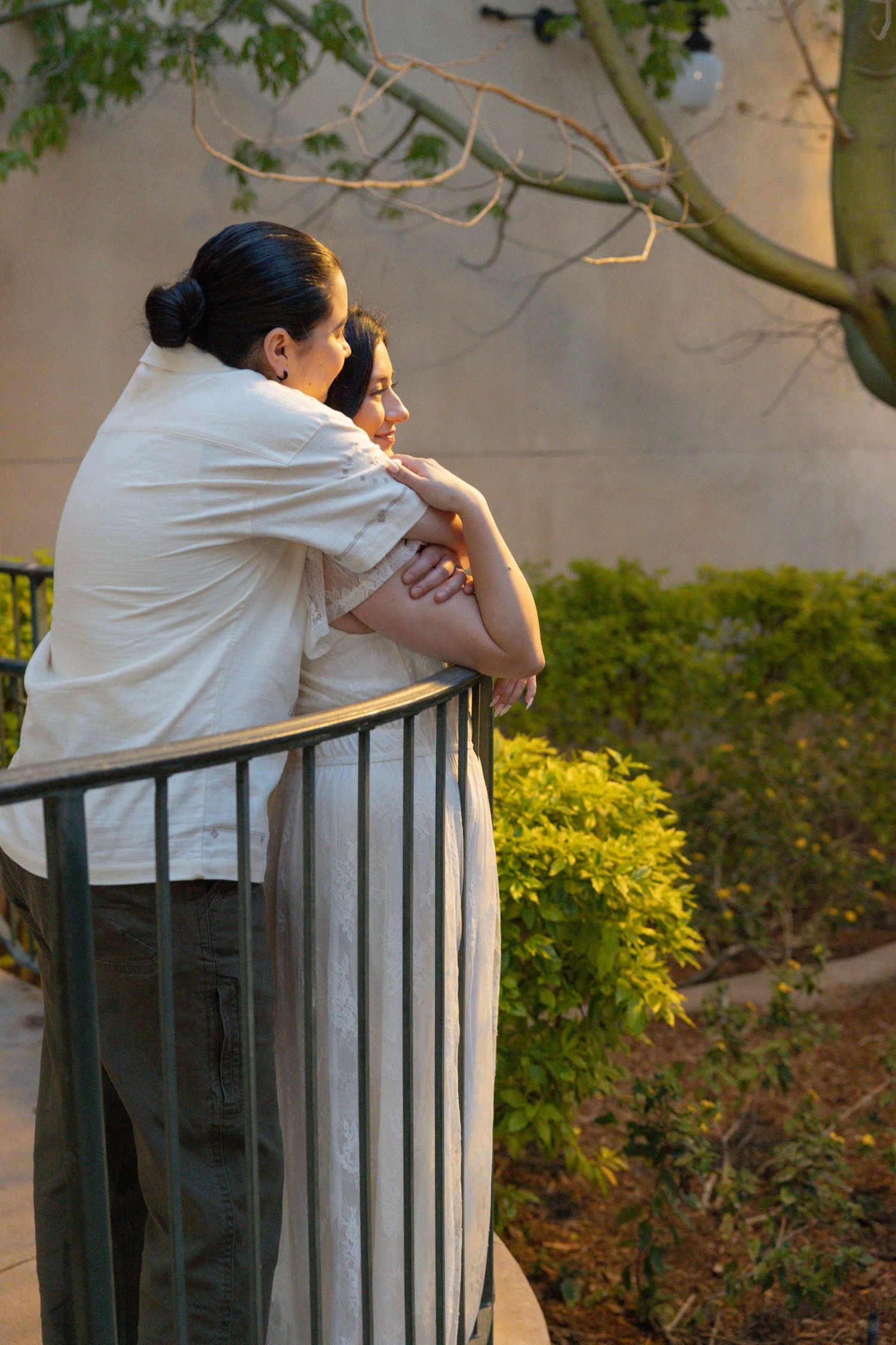 Two people lean on a railing overlooking a lush garden, sharing a tender embrace at golden hour. Couples Portraits San Diego California