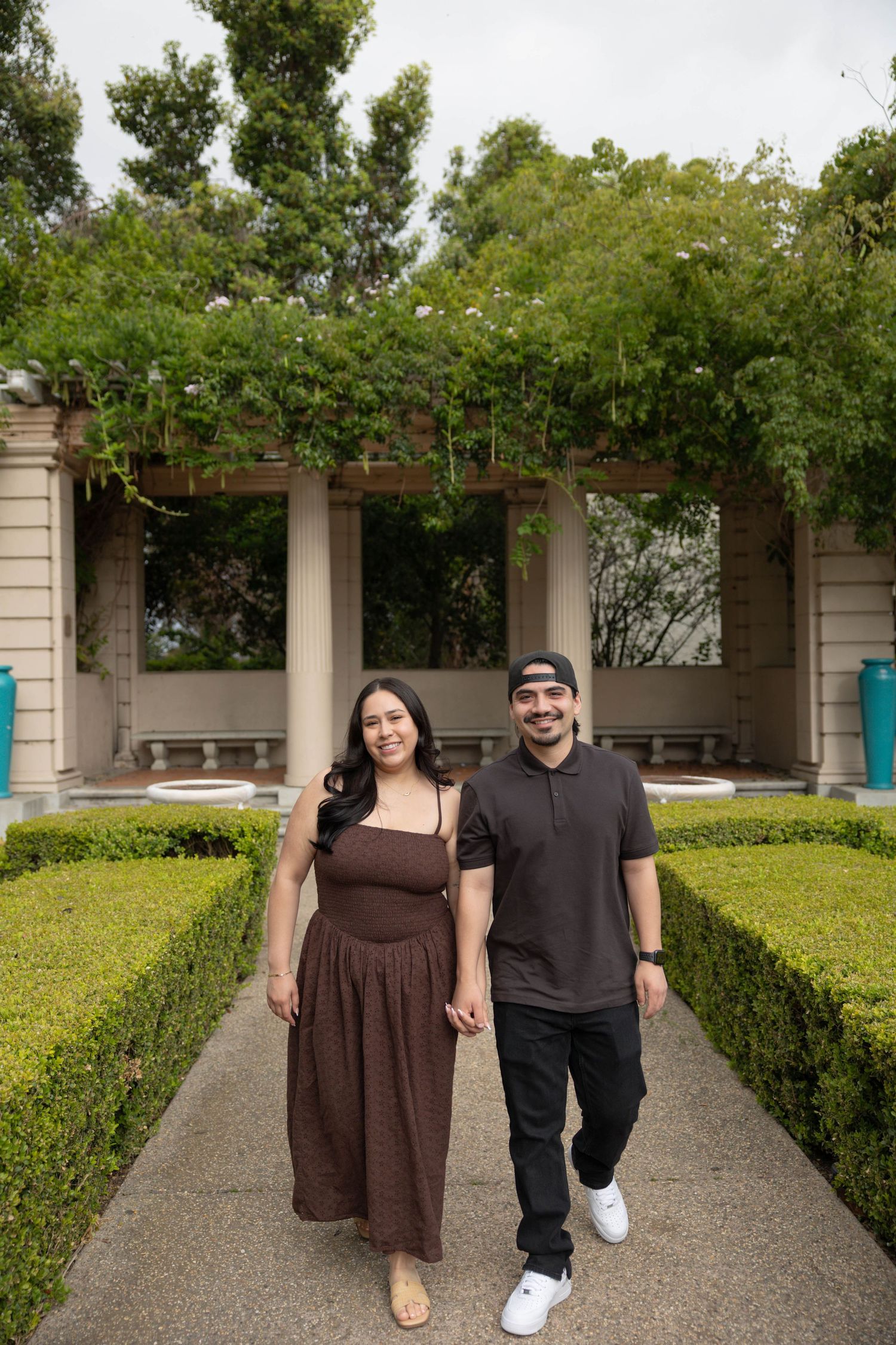 Couple holding hands walking along a garden path with trimmed hedges and vine-covered pergola columns. Couples Portraits San Diego Ca
