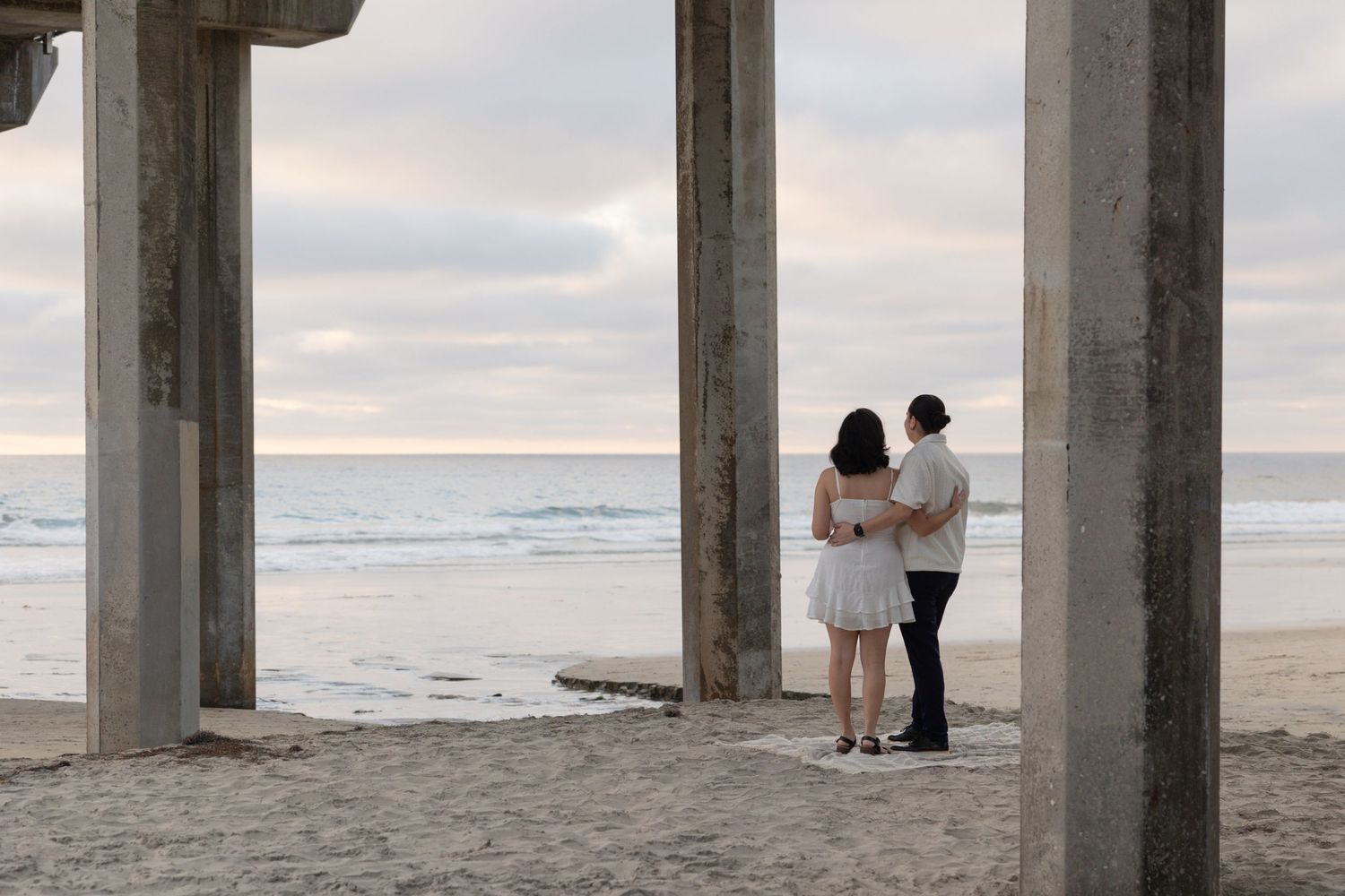 Couple standing together on sandy beach under concrete pier pillars at sunset with calm ocean in background.